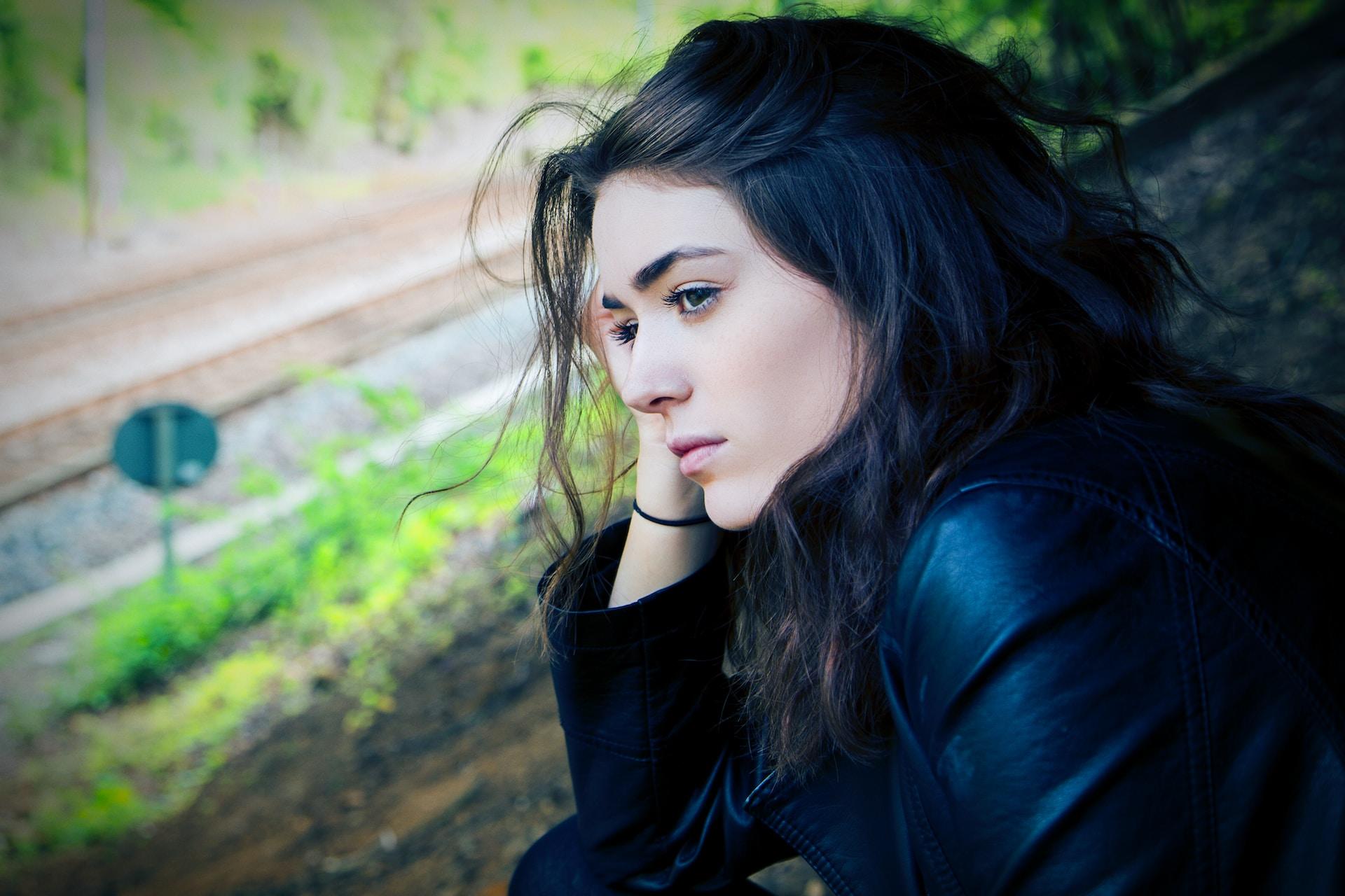 A woman wearing a black leader jacket sits outdoors, on a hill with railroad tracks at the bottom of the incline, holding her head in her hand as she gazes forlornly into the distance.