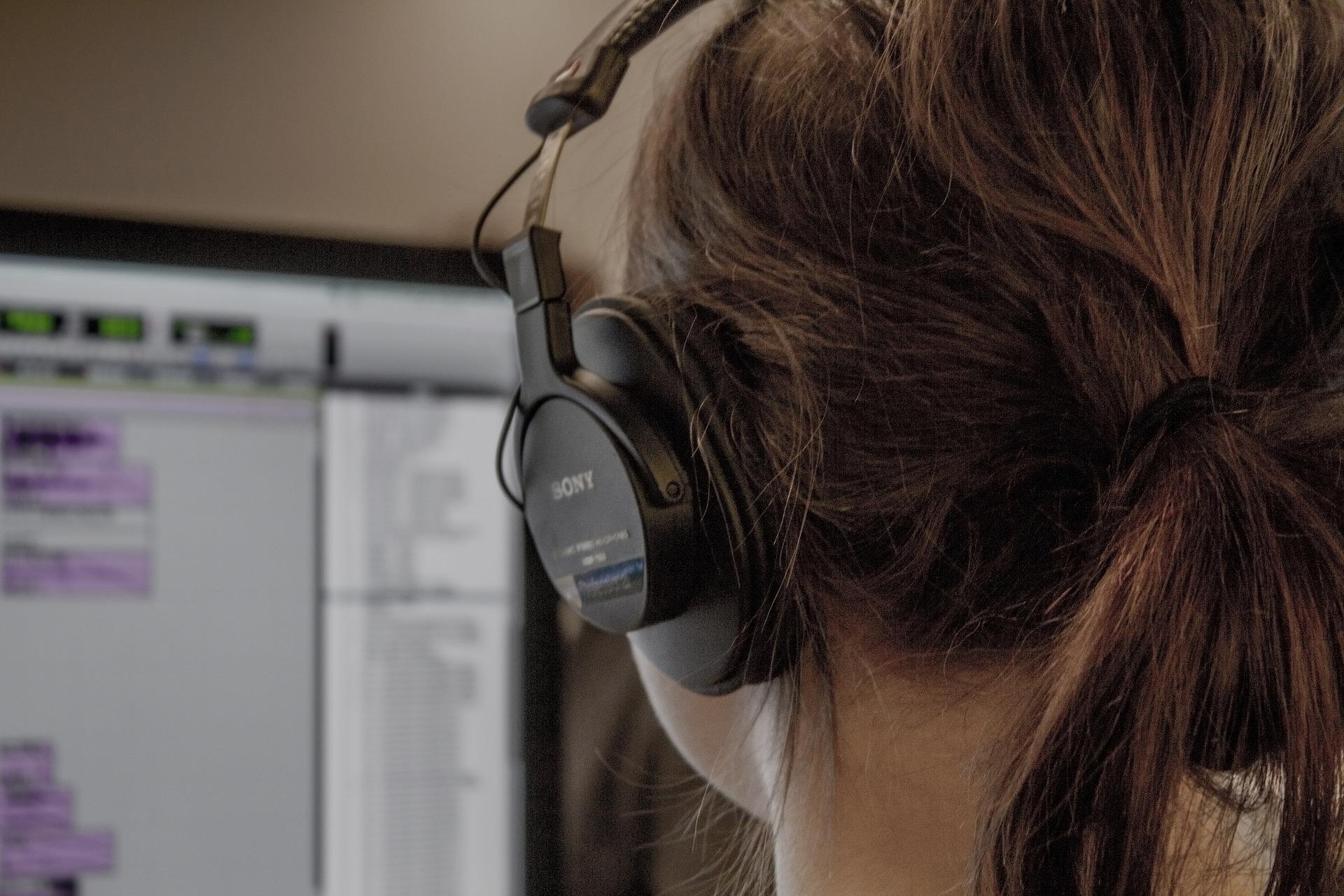 A person wearing headphones sits in front of a large computer screen which displays fields of text. Along the top of the screen is a row of buttons and down the right side is a hierarchical menu.,