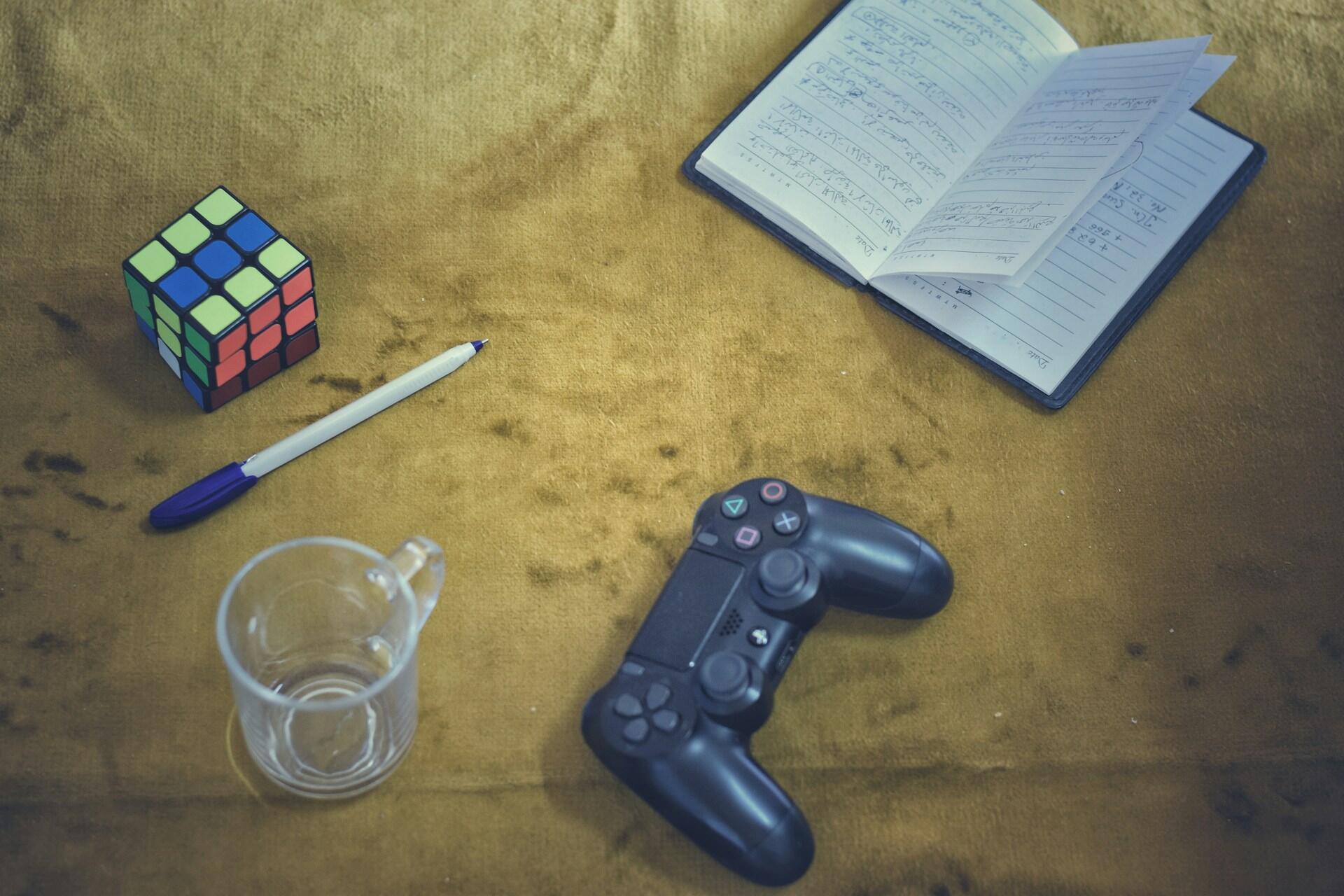 A colorful Rubik's cube, glass mug, pen, notebook with notes, and a black gaming controller on a mustard-colored surface.