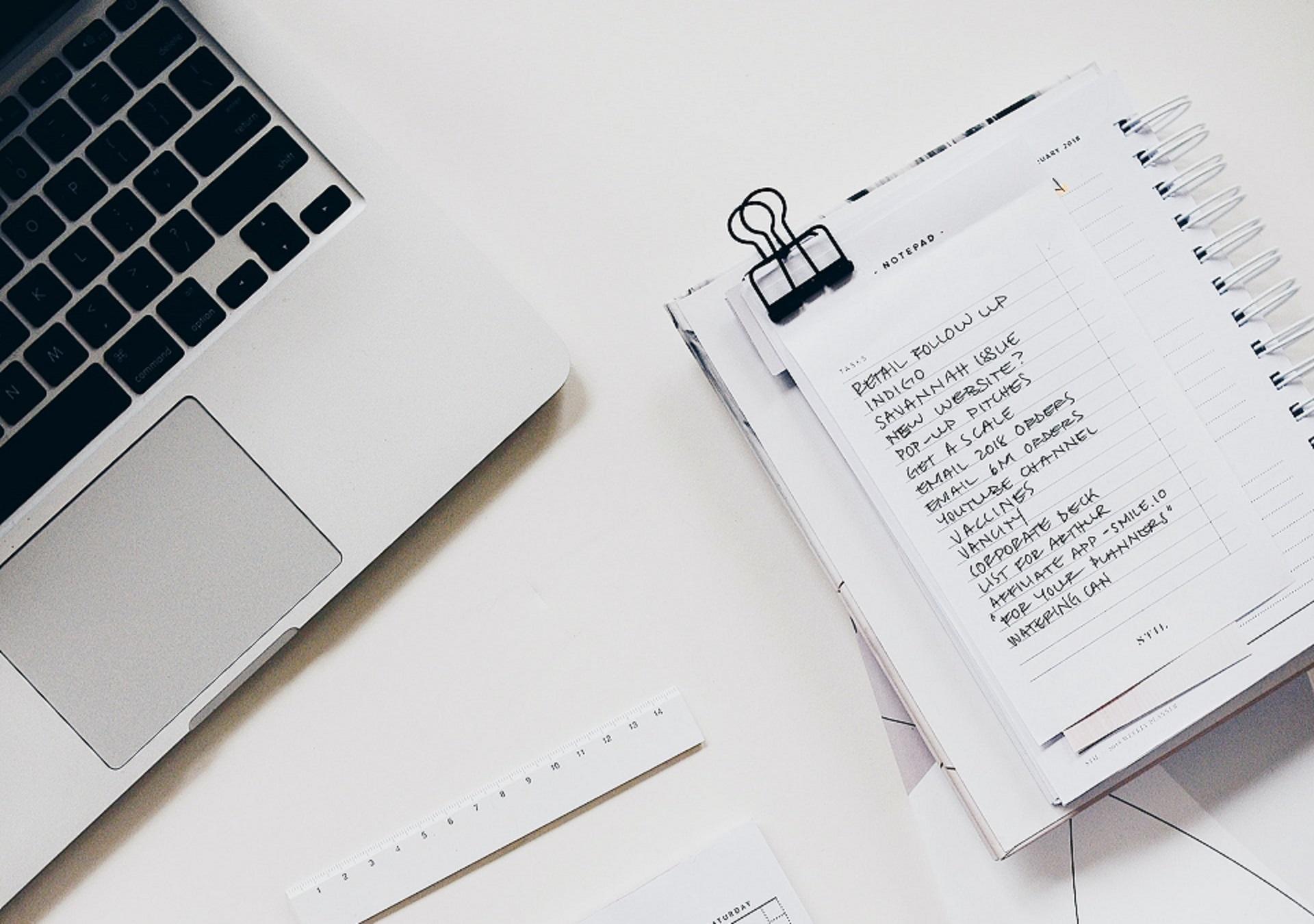 A notepad with a written list on it next to a laptop computer