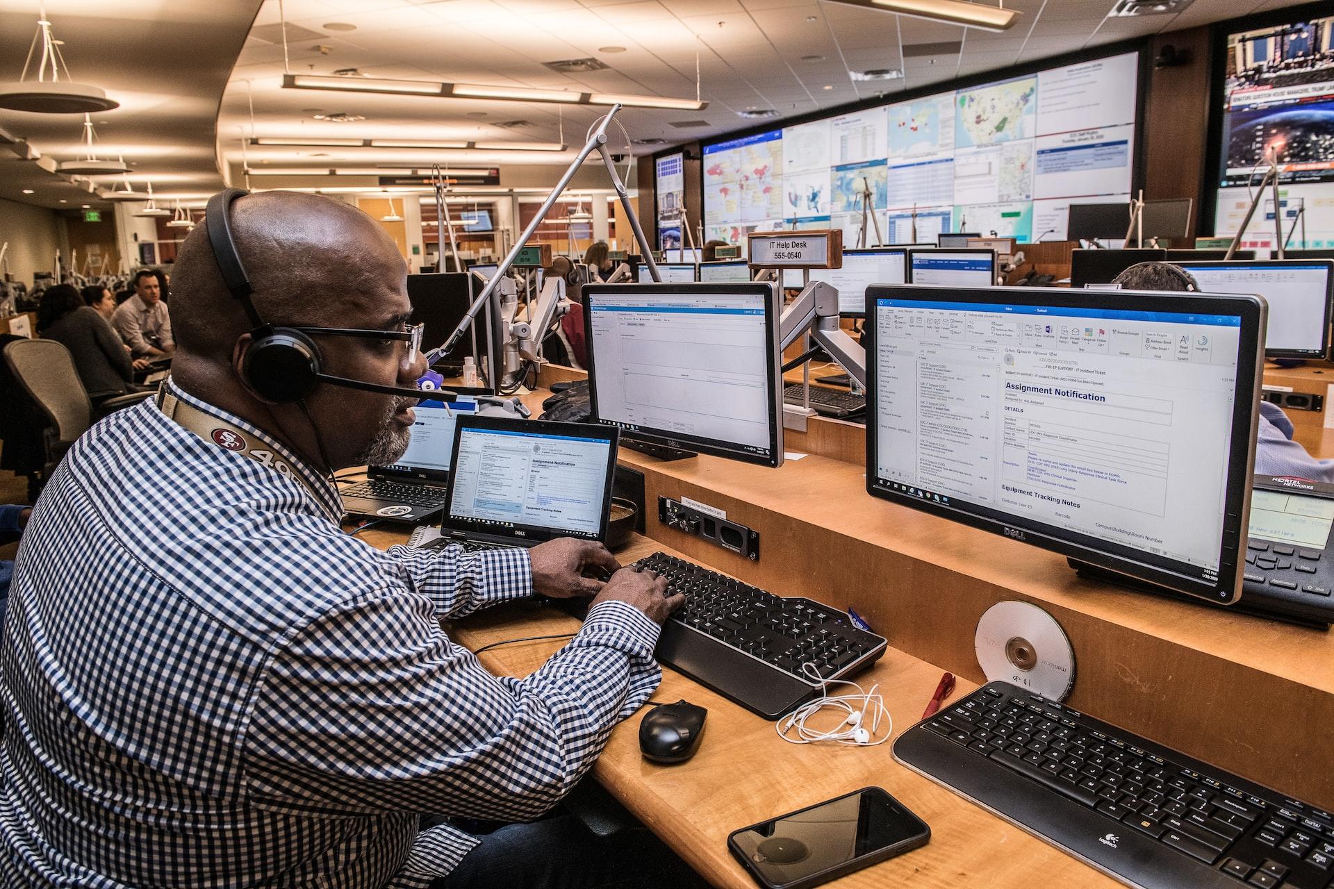 A man in a checkered shirt wearing a headset and sitting in front of a computer monitor