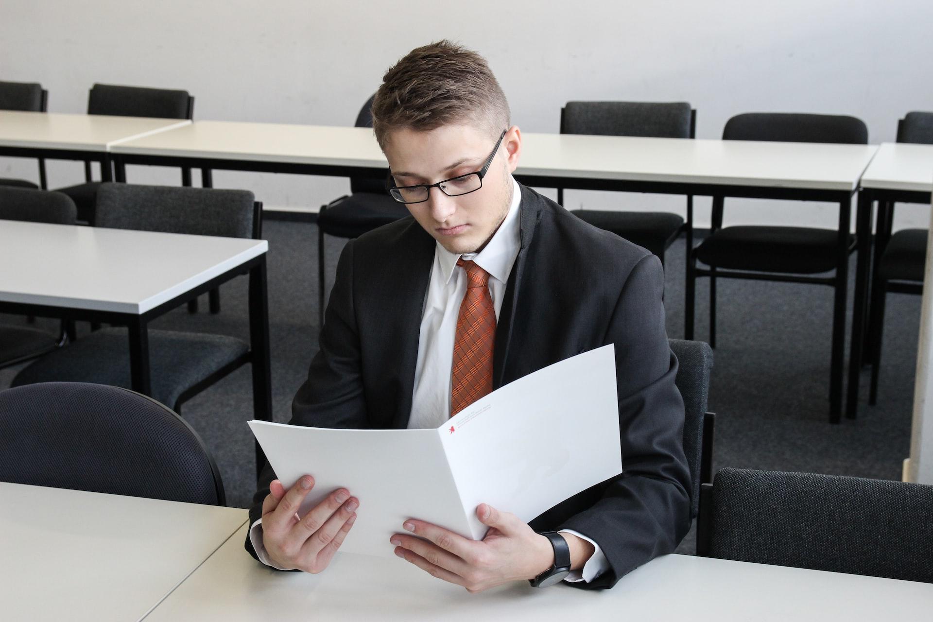 a man in a dark business suit and red tie reviews a document while sitting at a table