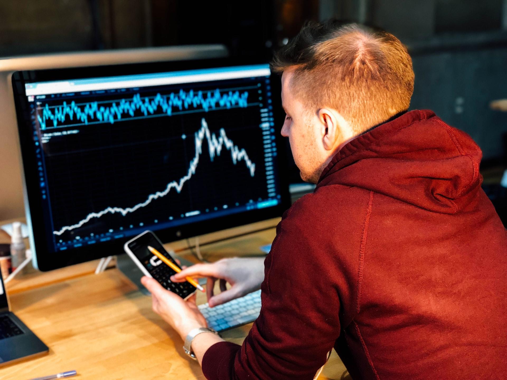 A red-haired man in a red hoodie sitting in front of a computer monitor with a mobile phone in his hand
