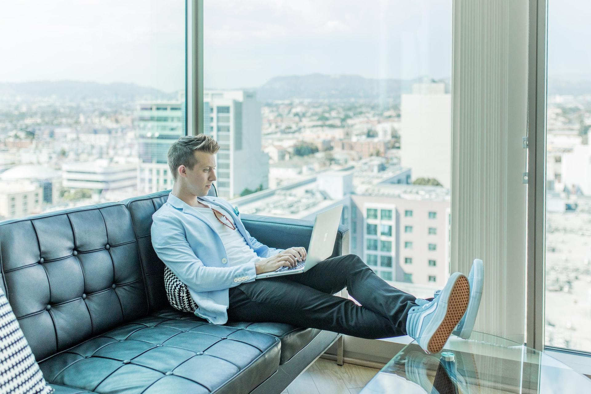 A man in a blue shirt on a couch, working on a laptop computer