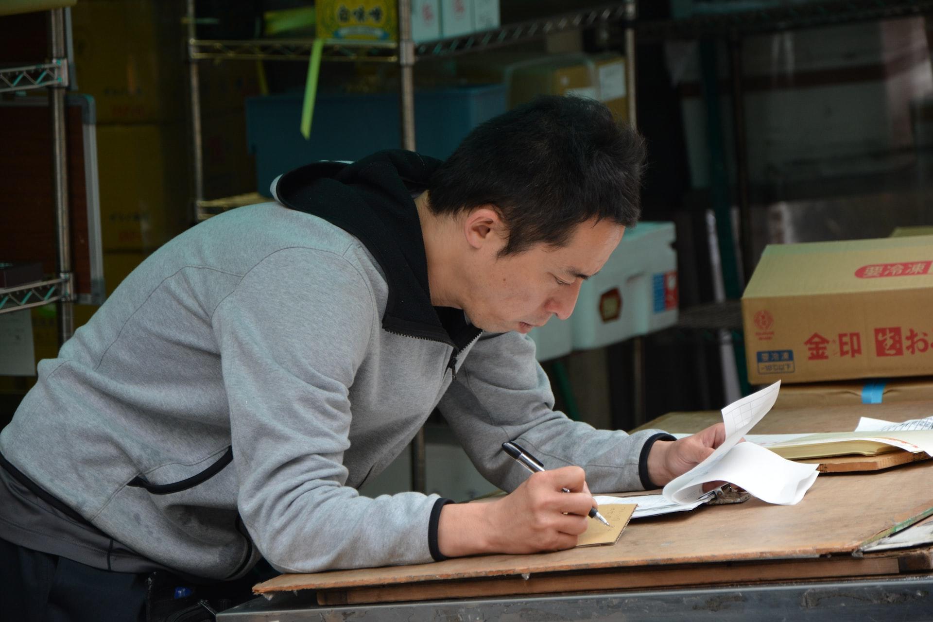 A man in a grey hoodie hunched over a countertop, writing on paper