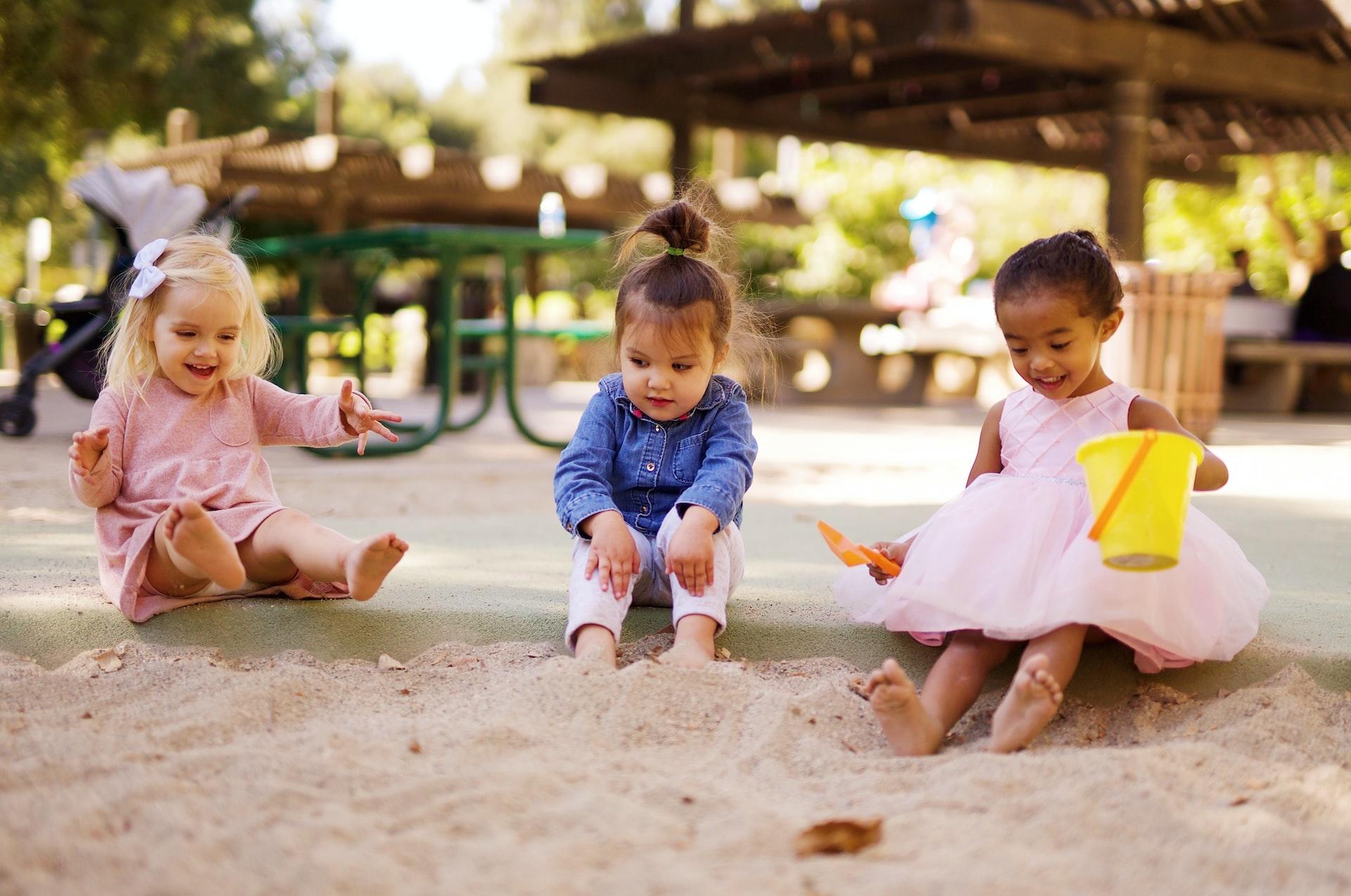 Three toddlers sitting at the edge of a sandbox with the one in the middle holding herself closed
