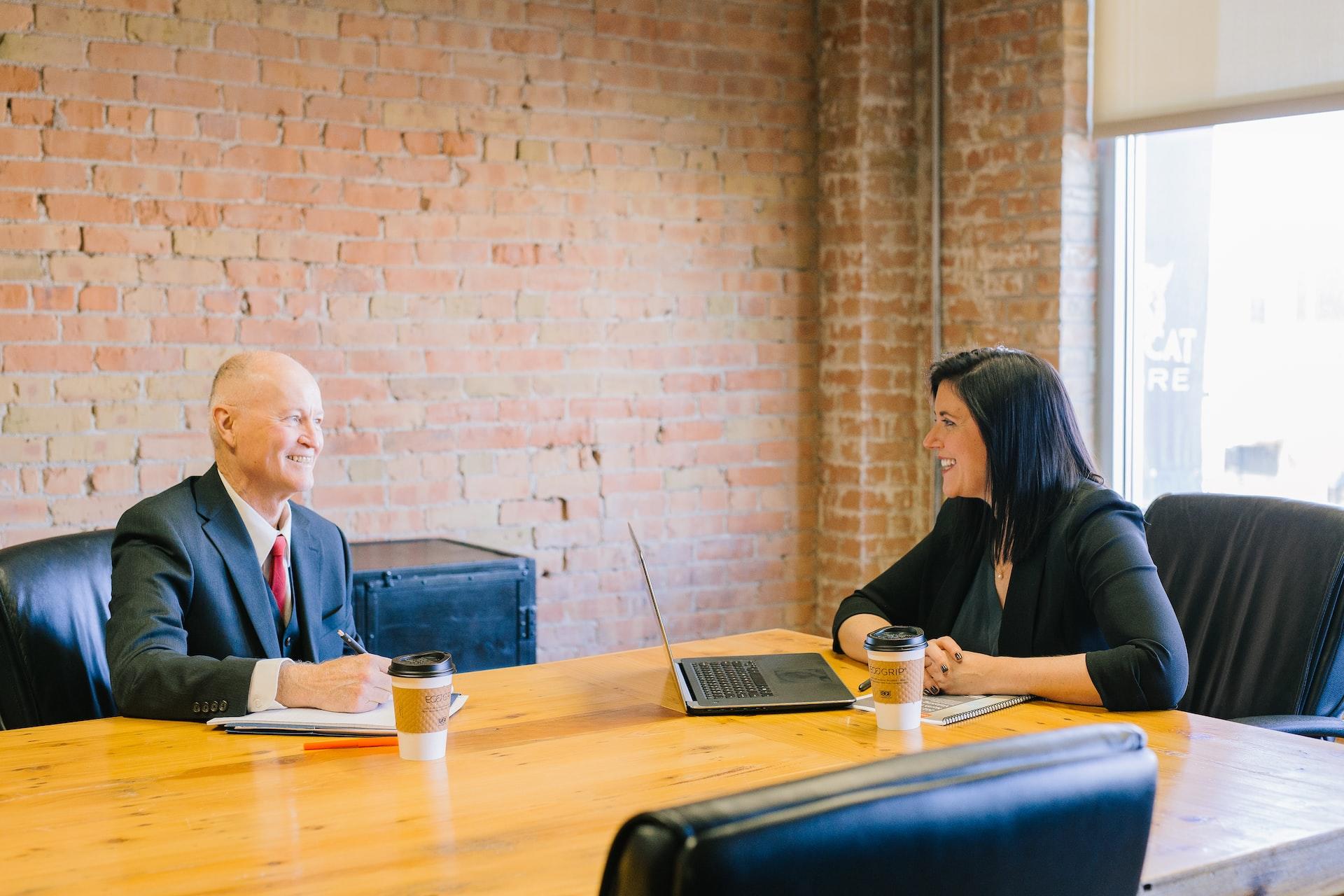 A man in a business suit and a woman in a dark coloured shirt seated at a large blond wood table with a brick wall in the background
