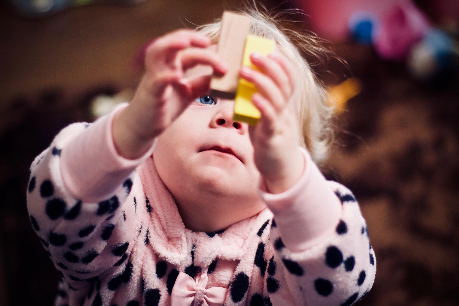 A blond toddler in a pink and black jacket holds two wooden blocks in the air and looks at them