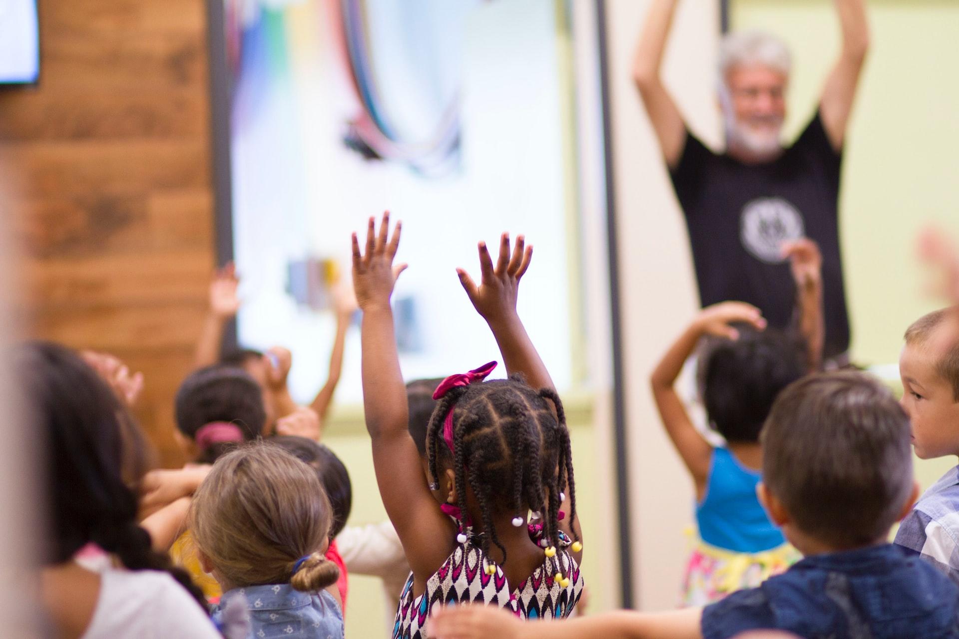 A group of preschool students with their arms raised, copying their teacher's movements