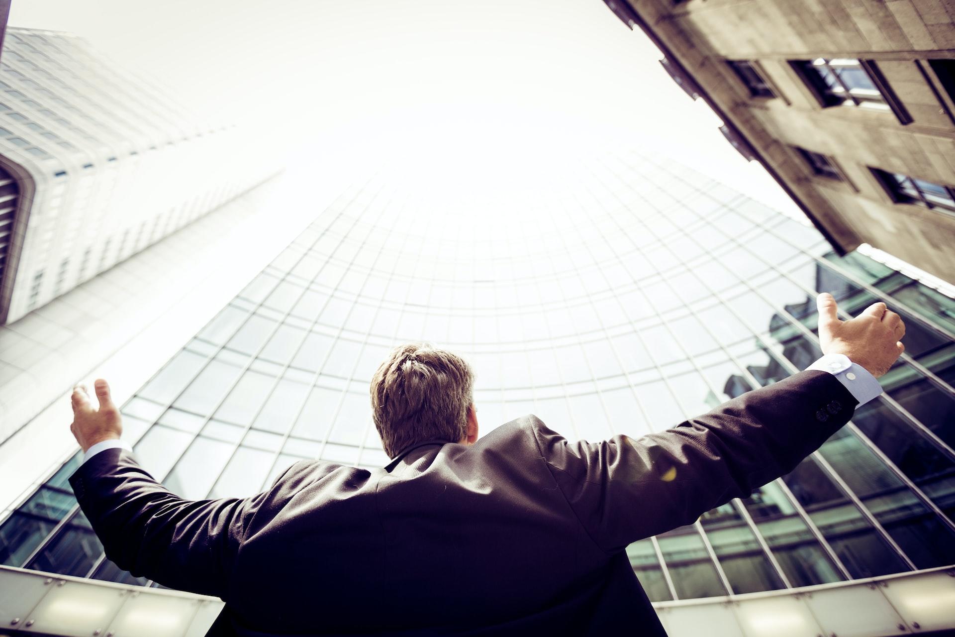 A man in a business suit standing with arms raised in front of a glass-fronted building with an older building to the side