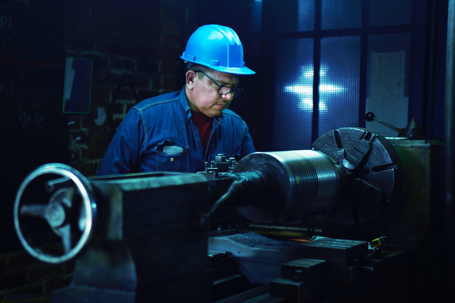 A man in a blue hardhat and blue coveralls working a lathe. 