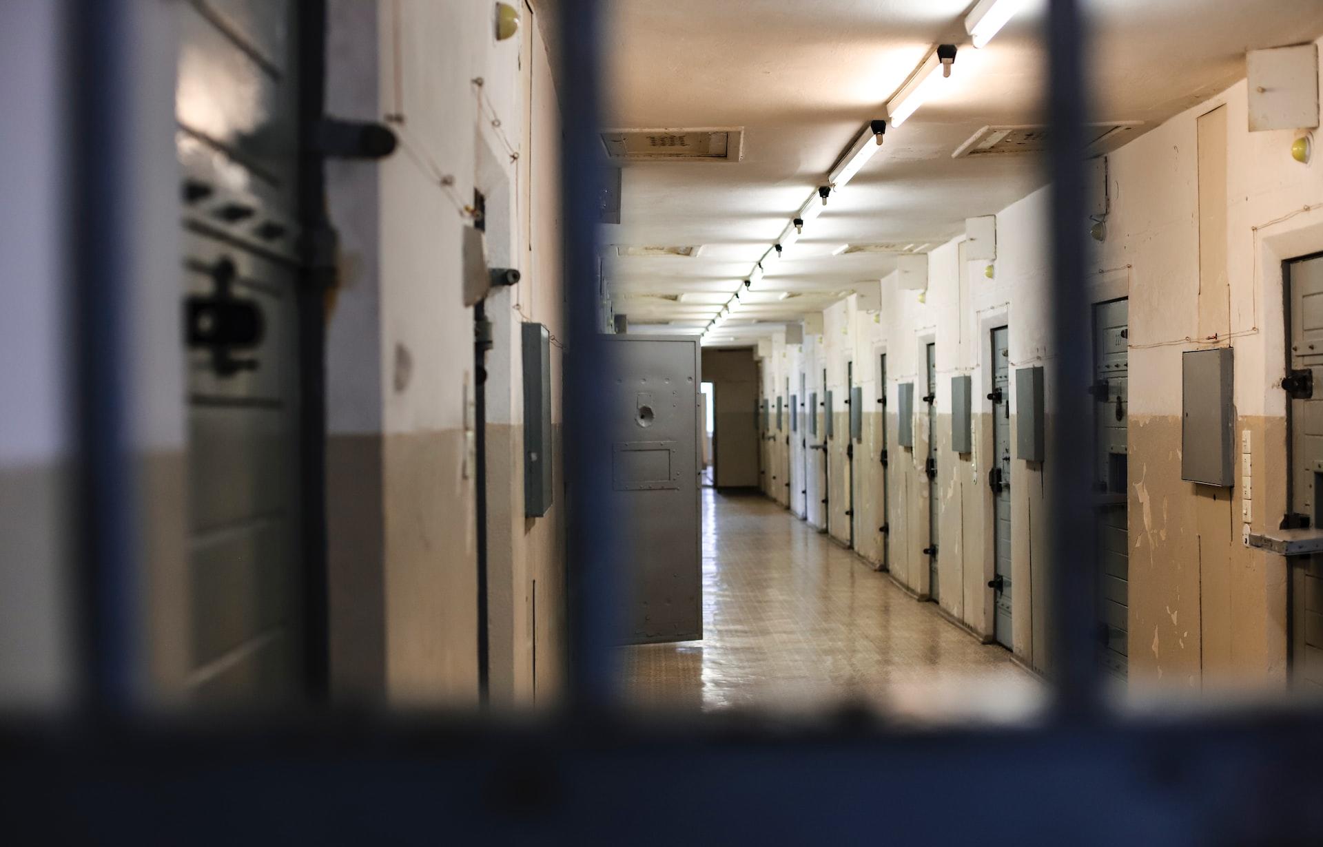 A prison cellblock corridor painted ochre seen through bars with one grey cell door open in the distance.