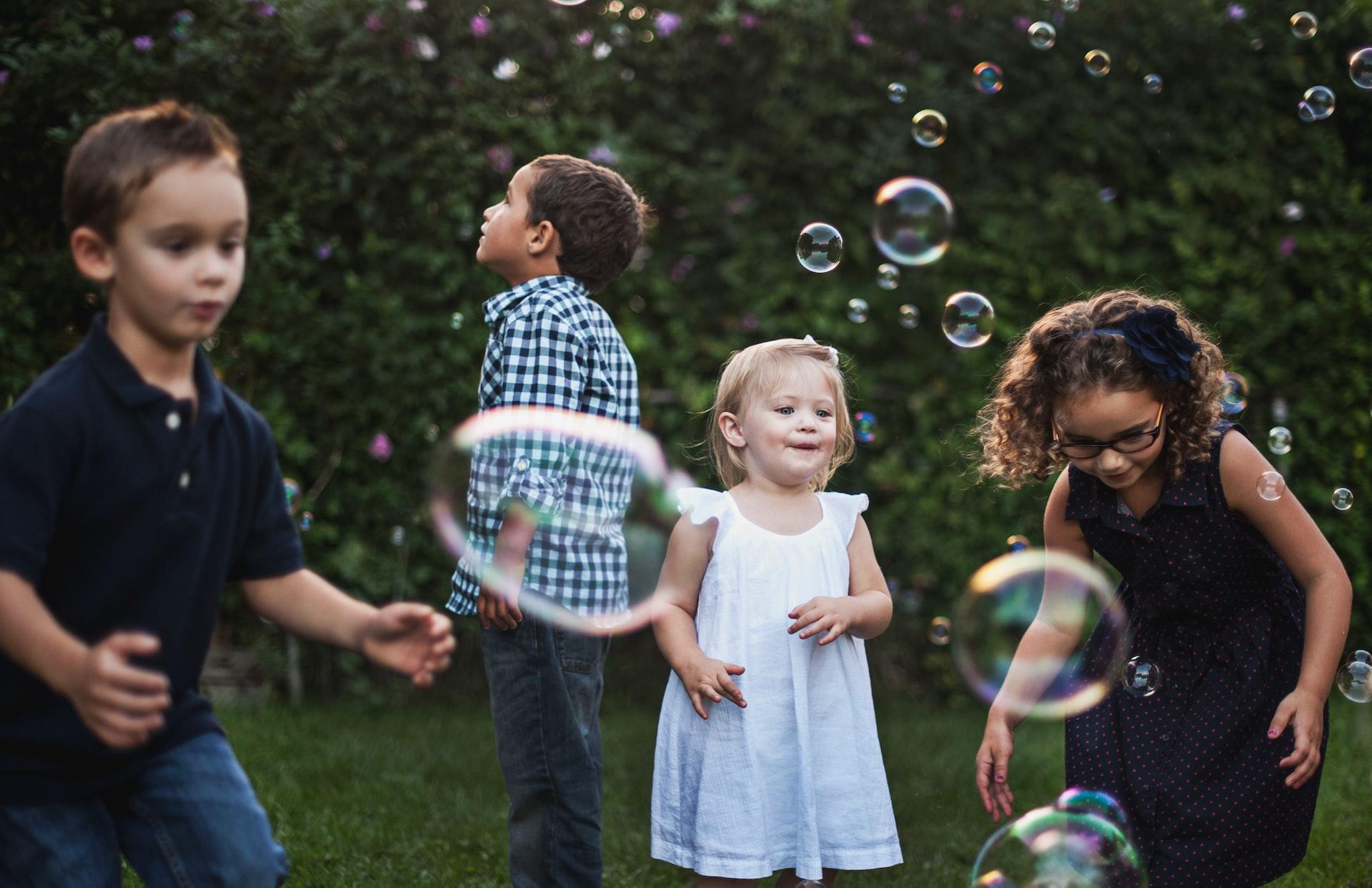 A group of children chasing after soap bubbles in the park while a small girl in a light blue dress observes them