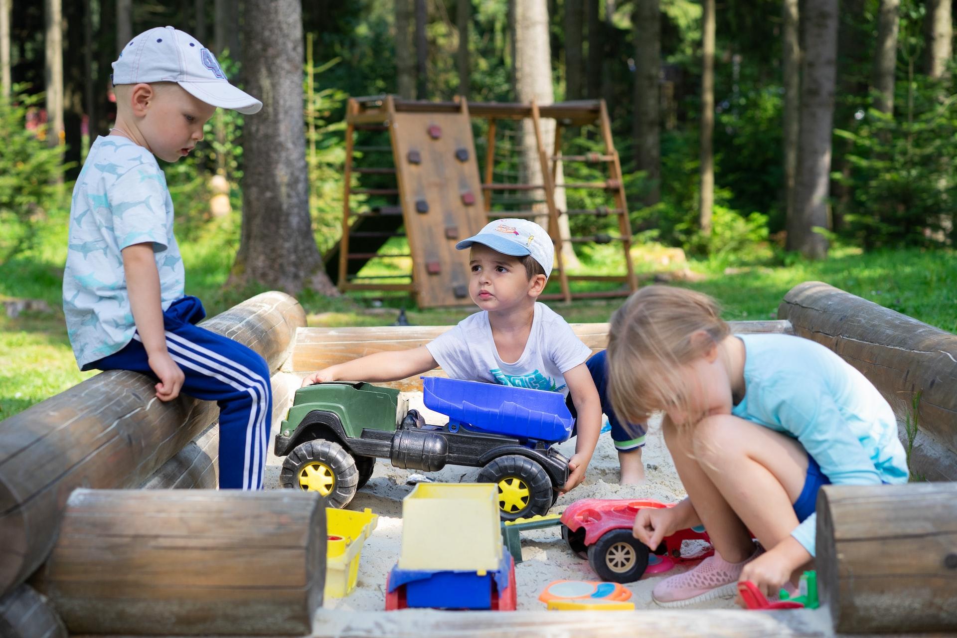 Three young boys in a sandbox bordered by logs playing with toy construction trucks