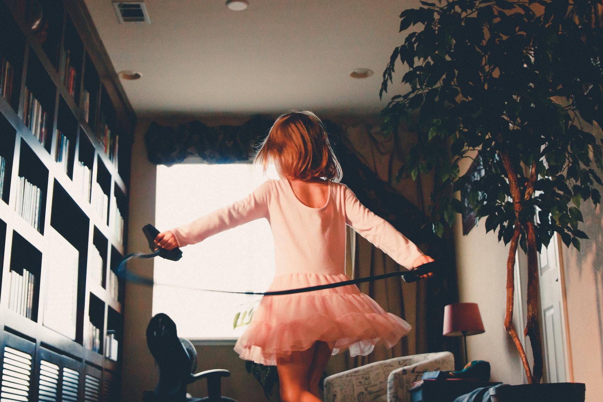 A young girl in a ballet pink tutu, holding a black ribbon in both hands, dances through a library. 