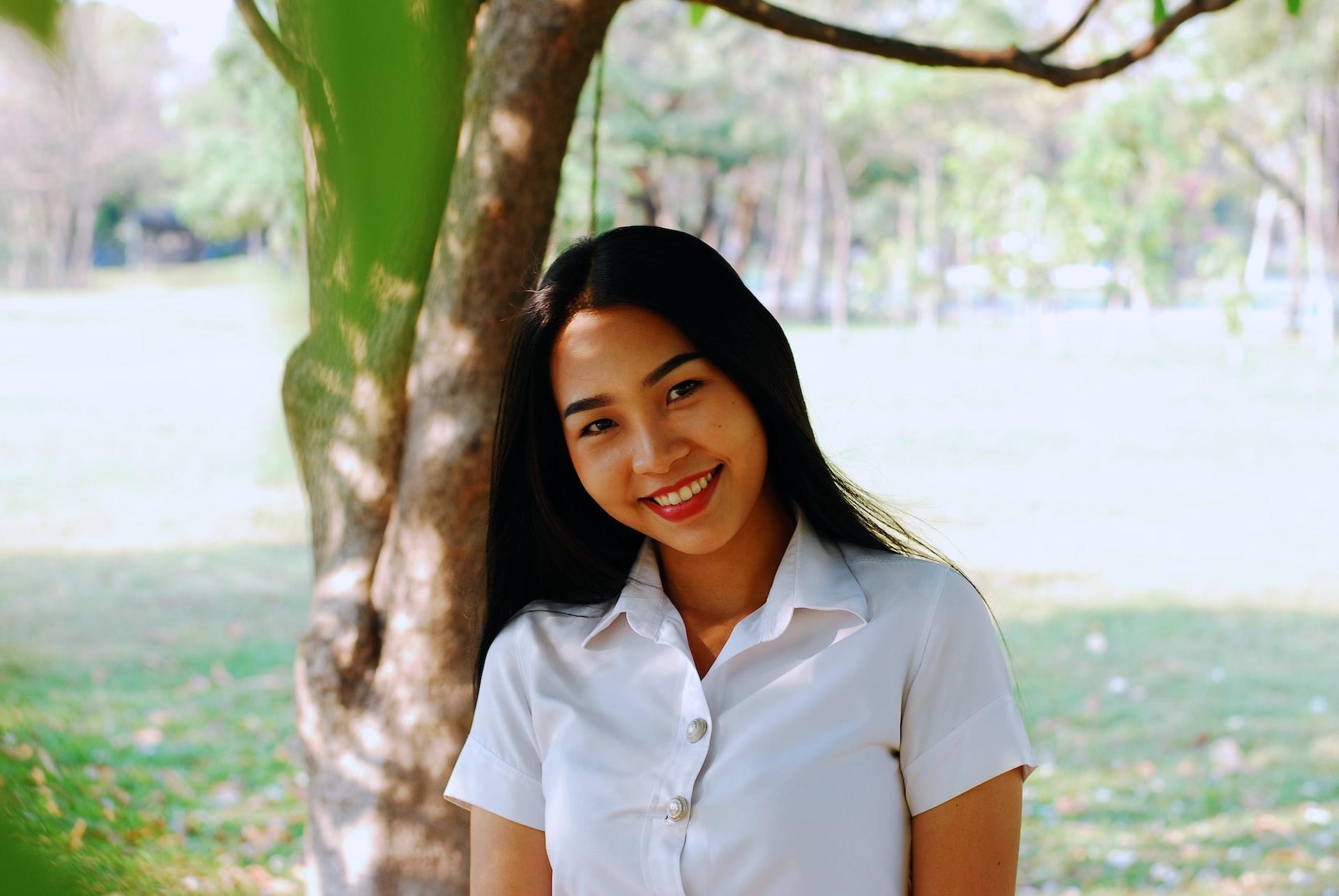 A young woman wearing a white blouse leans against a tree, smiling, on a sunny day.