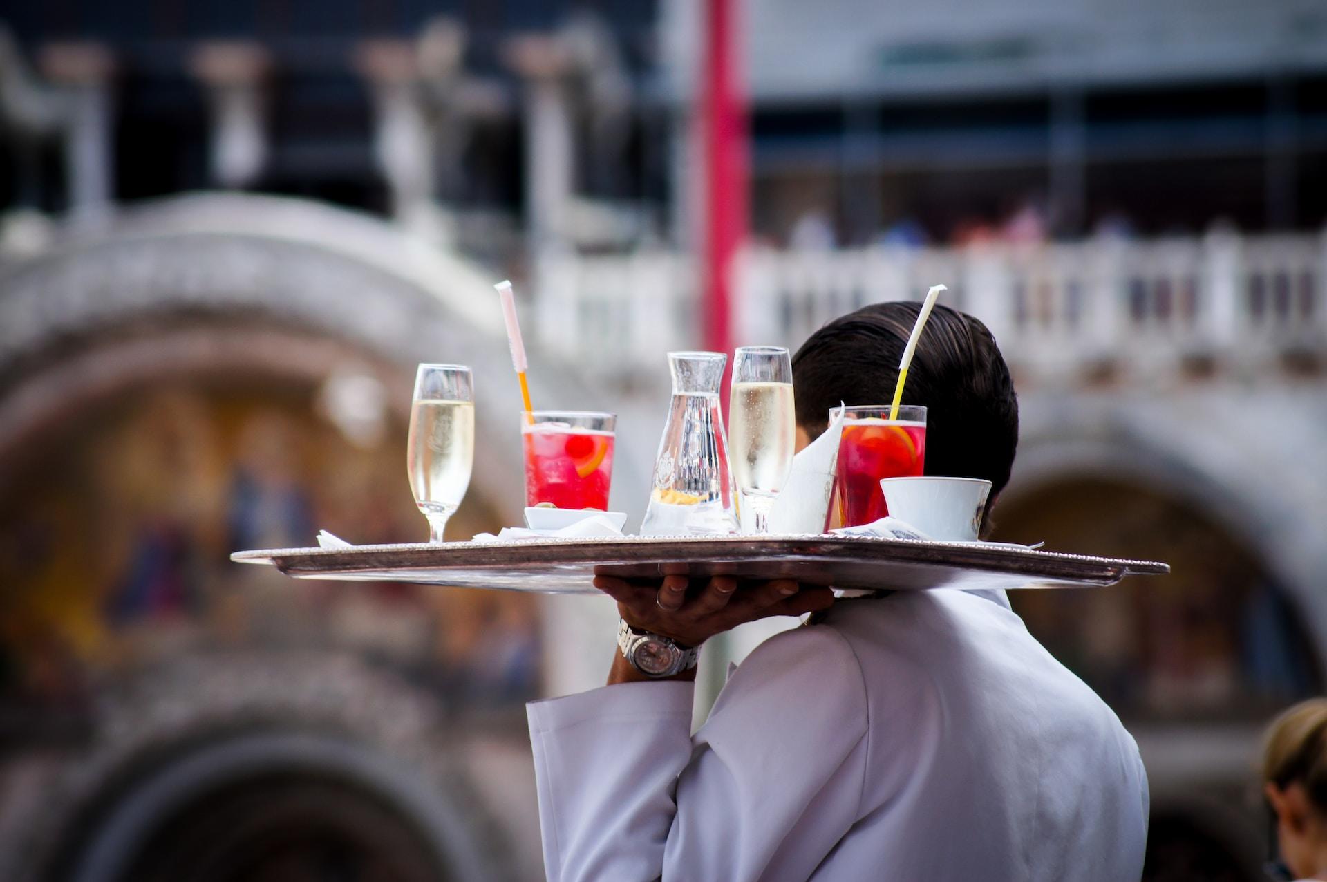 A restaurant server wearing a white shirt carries a tray laden with red-coloured drinks at shoulder-level. 
