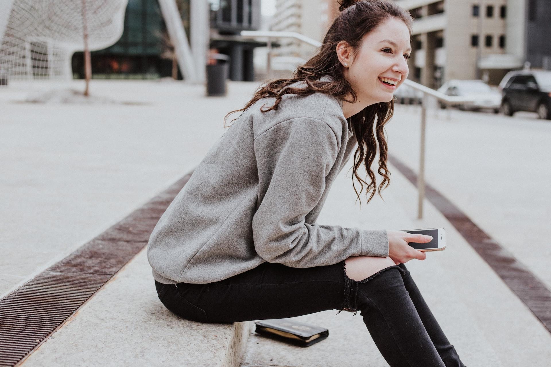 A young woman in a grey jumper and ripped black jeans leans forward while sitting on a concrete step, smiling. 