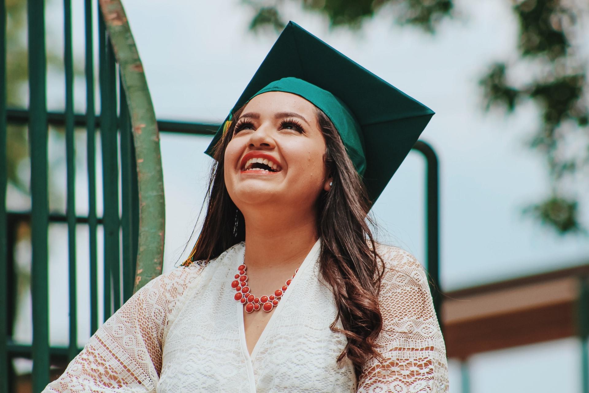 A woman wearing a white lacy top and a green graduation mortarboard smiles while looking up. 