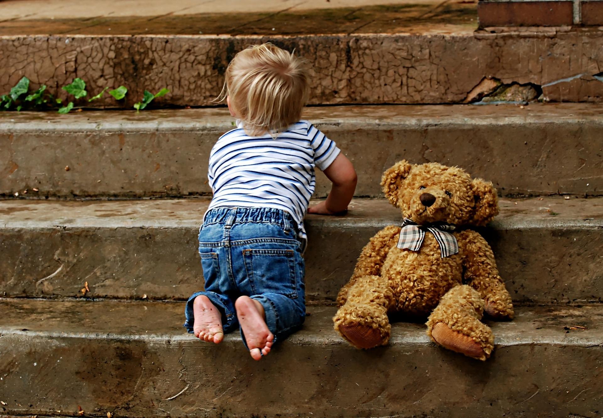 A toddler climbing up the stairs with a teddy bear on his side.