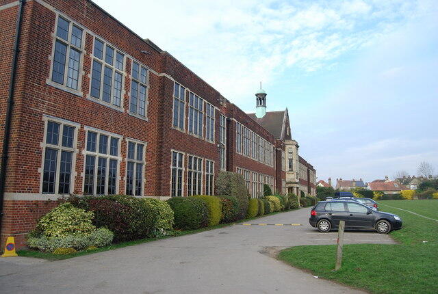 Side view of a brick school building with large windows, lush greenery, and a parking area, set against a cloudy sky.