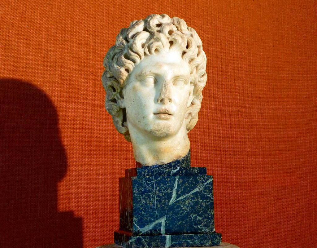 A marble bust with curly hair rests on a dark marble pedestal against an orange textured background, casting a soft shadow.