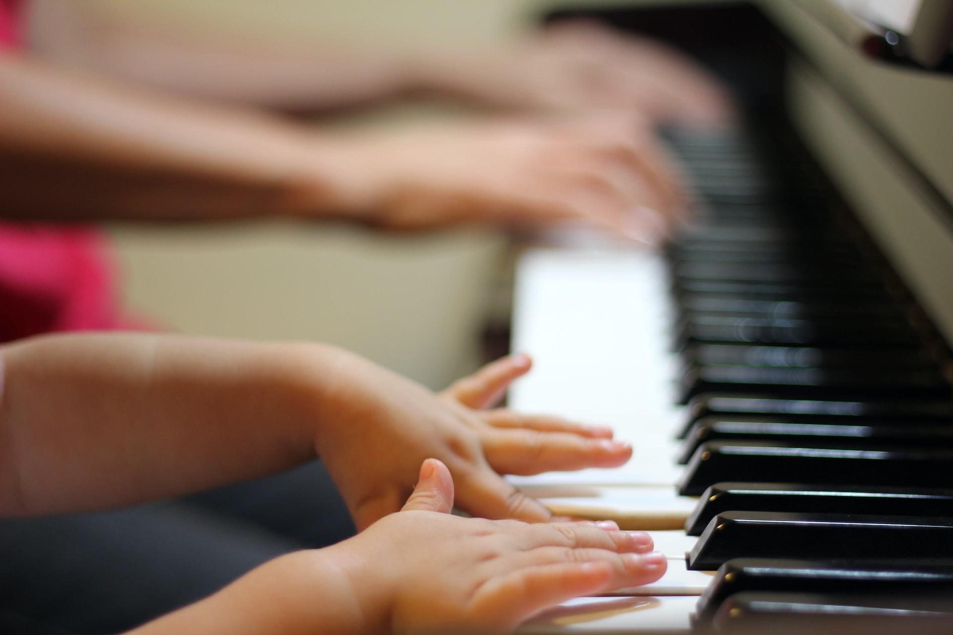 A closeup of a piano keyboard seen from the treble end with a child's pair of hands in the foreground and adult hands in the background.