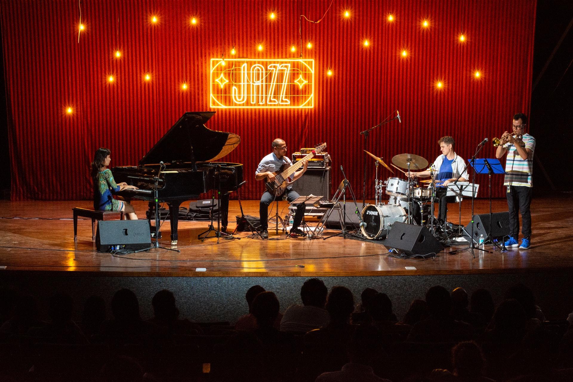 A jazz combo featuring a grand piano and other instruments on a stage backlit by a red curtain with the word jazz emblazoned in golden lights.