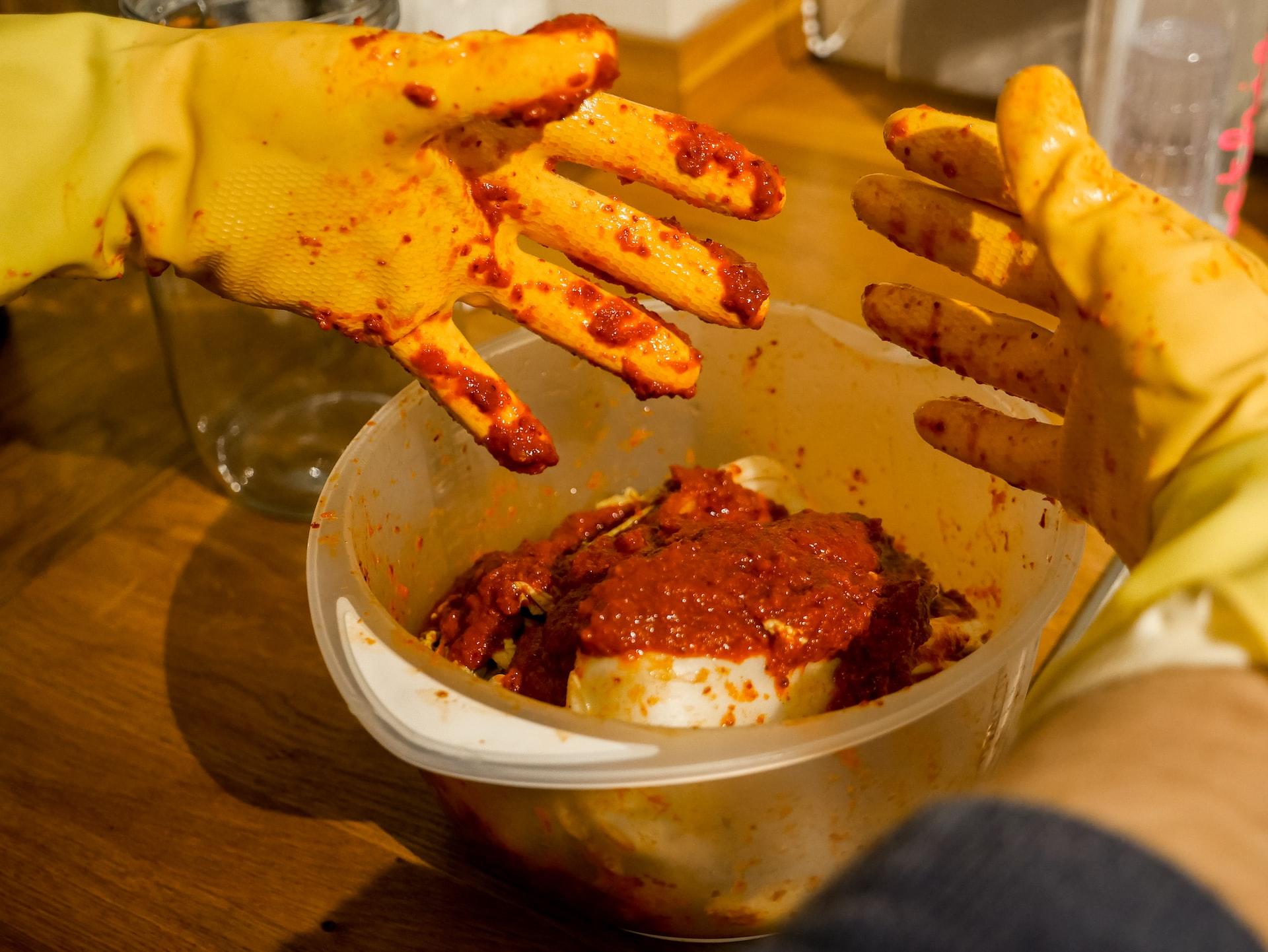 A person wearing yellow cleaning gloves mixes reddish sauce and seasonings into a bowl of cabbage to make kimchi.