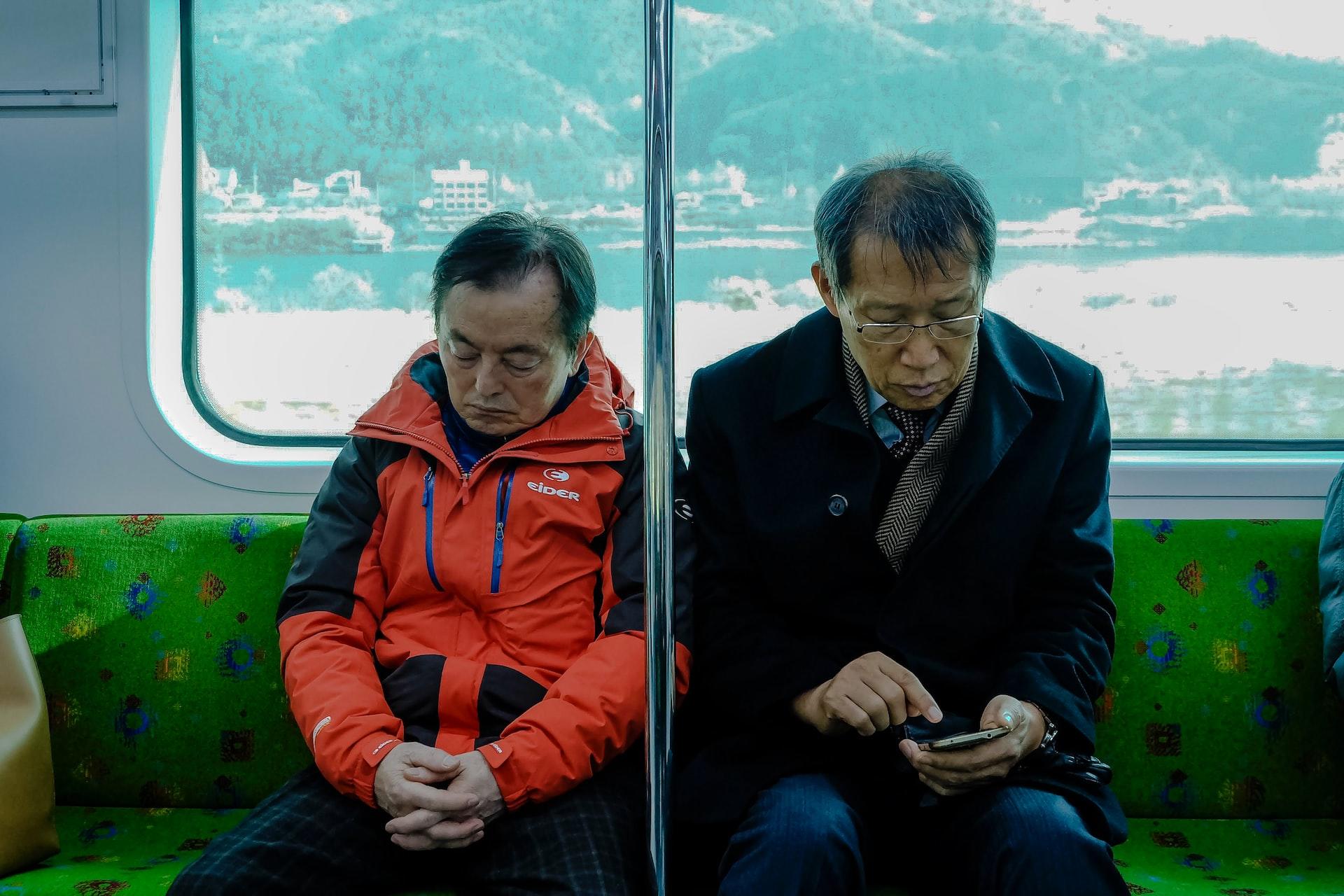 One old man in an orange jacket snoozes on the train next to another old man wearing a dark jacket and glasses who looks at his phone.