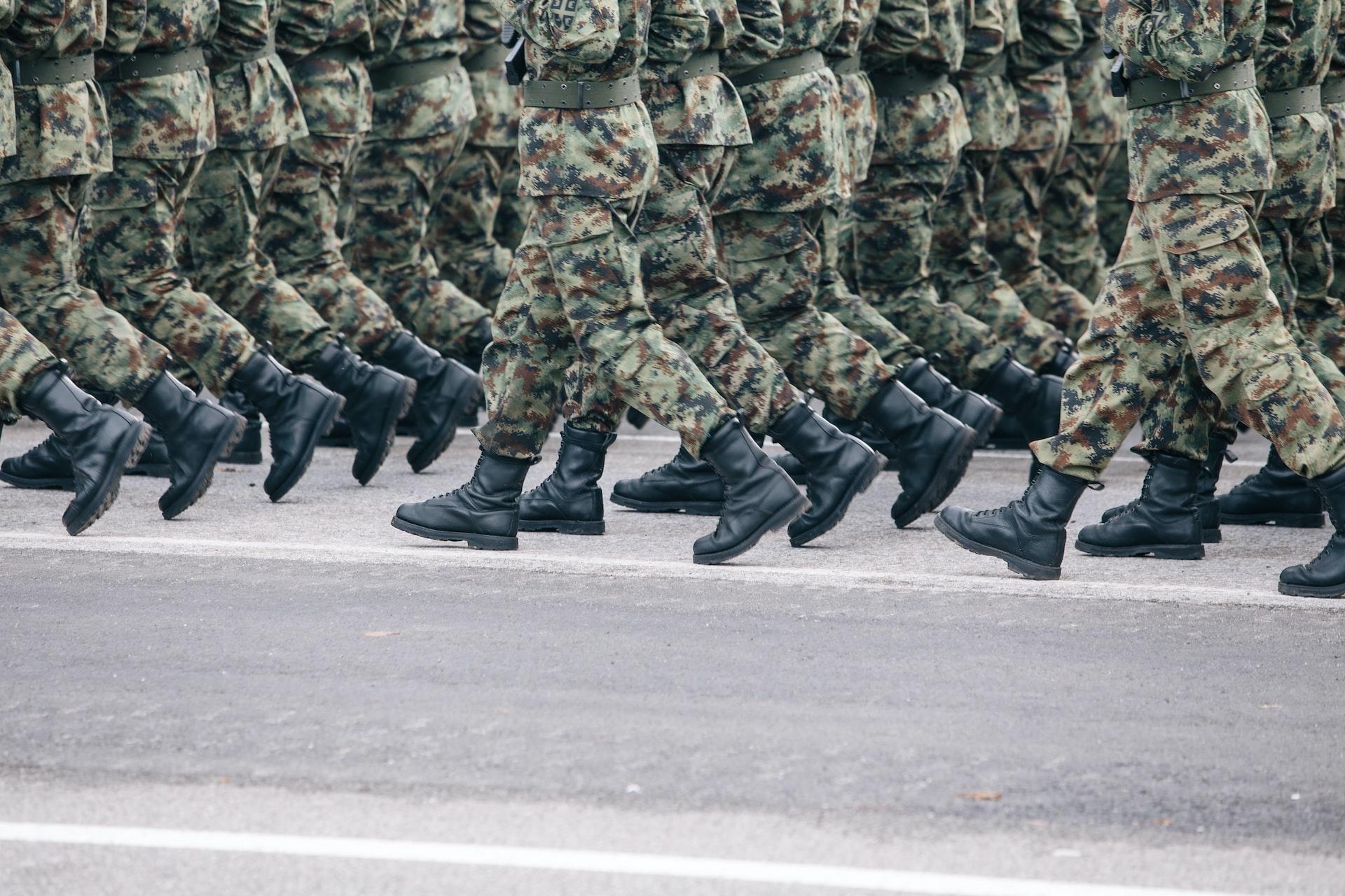 A platoon of soldiers wearing camouflage uniforms and web belts marches in formation. 