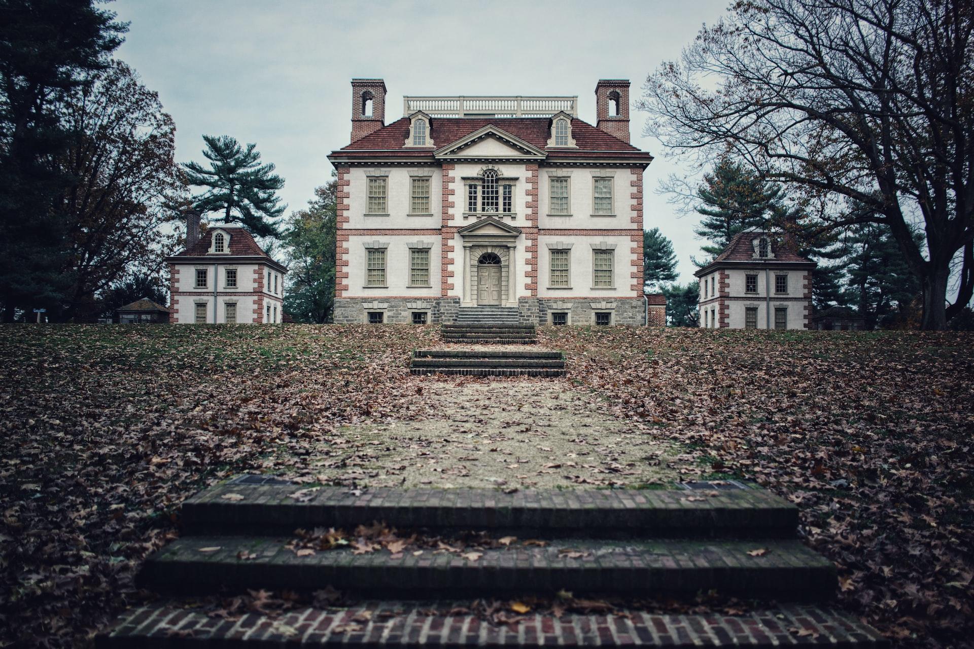 An eerie-looking two-storey house flanked by two smaller buildings at the end of a very long, leaf-strewn walkway which features occasional steps.