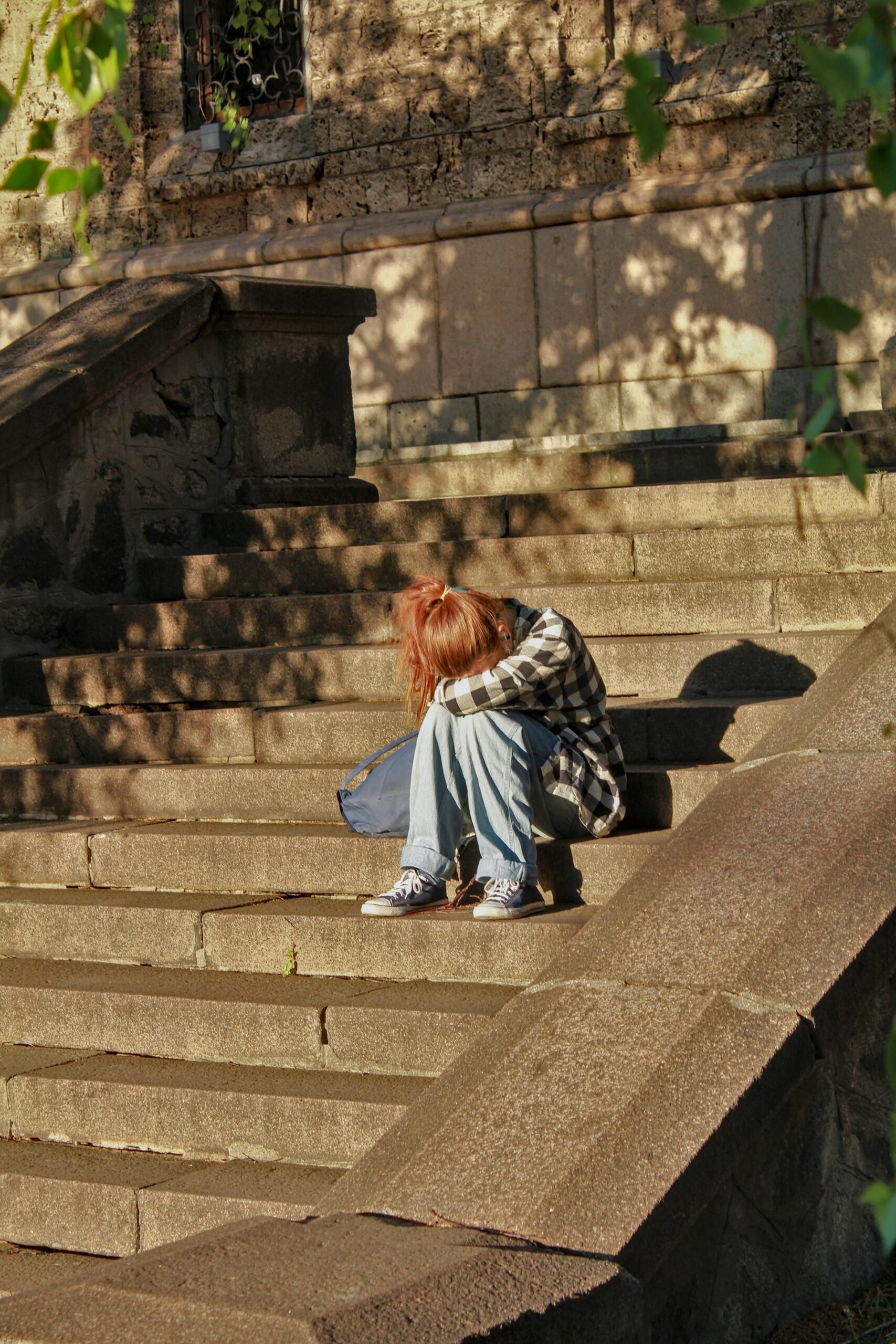 A person in jeans and a plaid shirt sits on stone stairs with their head on their knees.