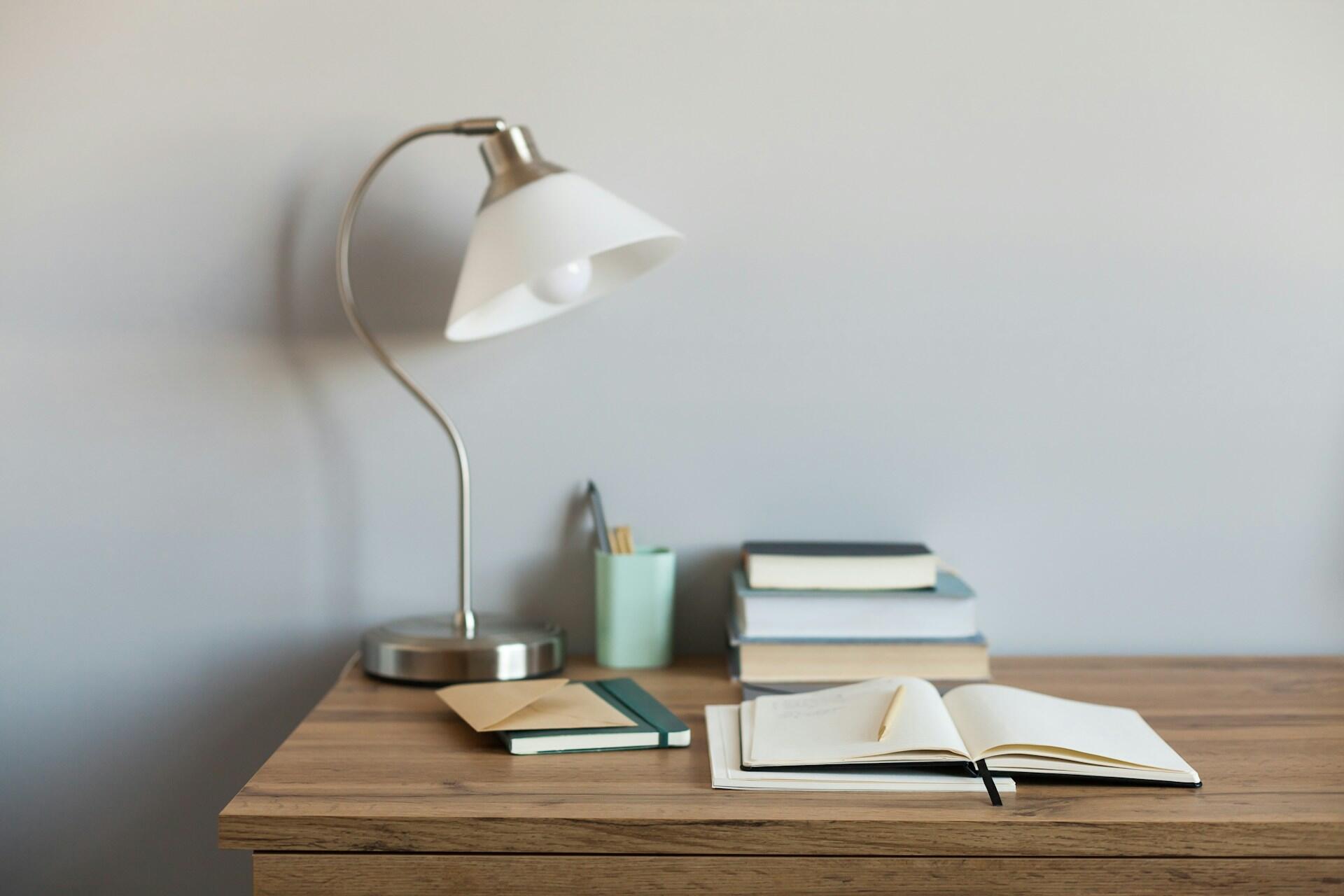 A desk with a stack of books, an open notebook and a lamp.