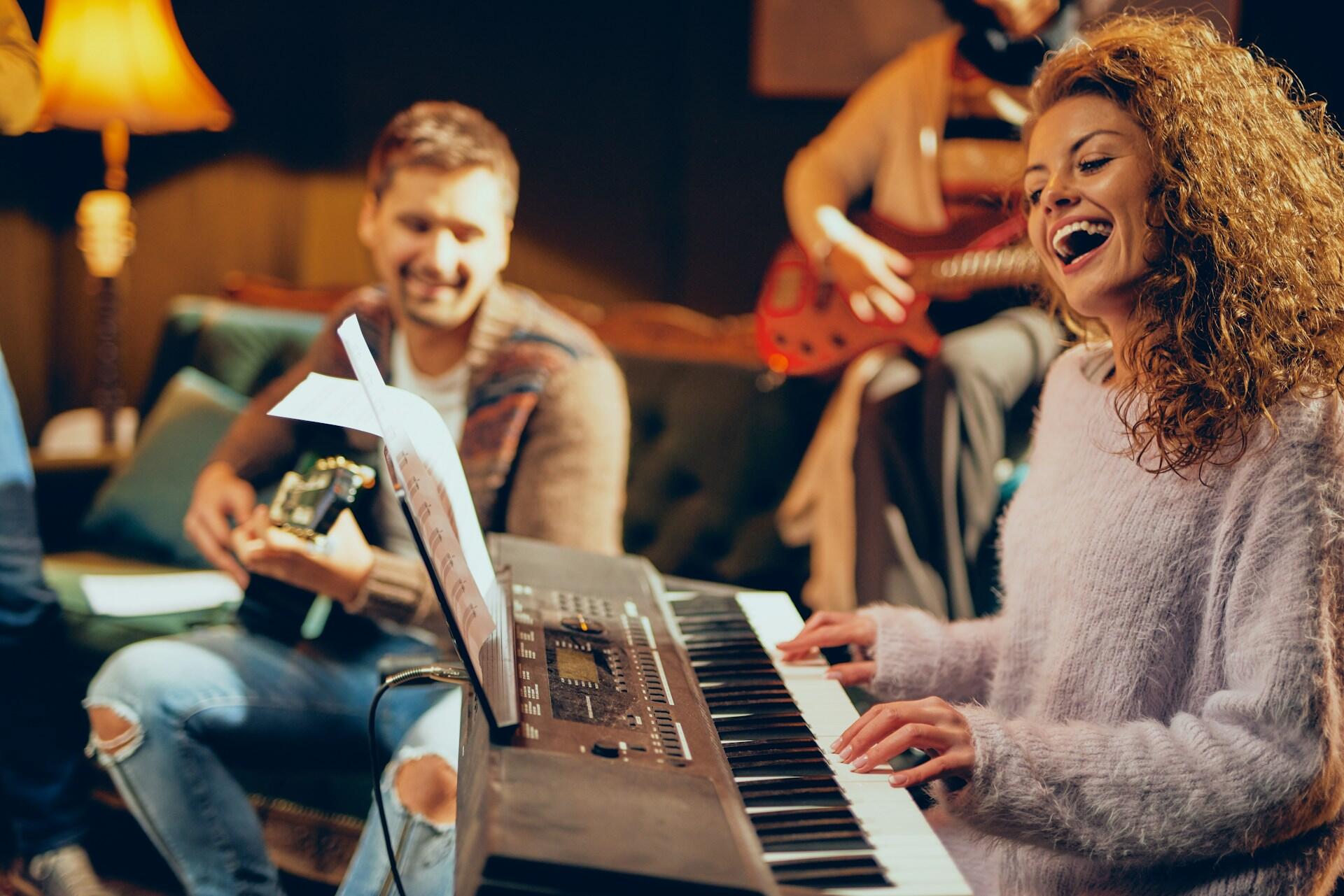 Group of musicians in a cozy room playing keyboard, guitar, and bass with sheet music on a stand.