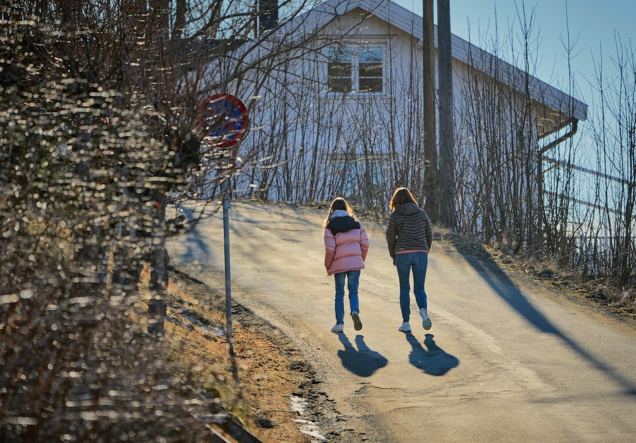 Two kids walking uphill on a sunlit pathway near a house and bare trees.