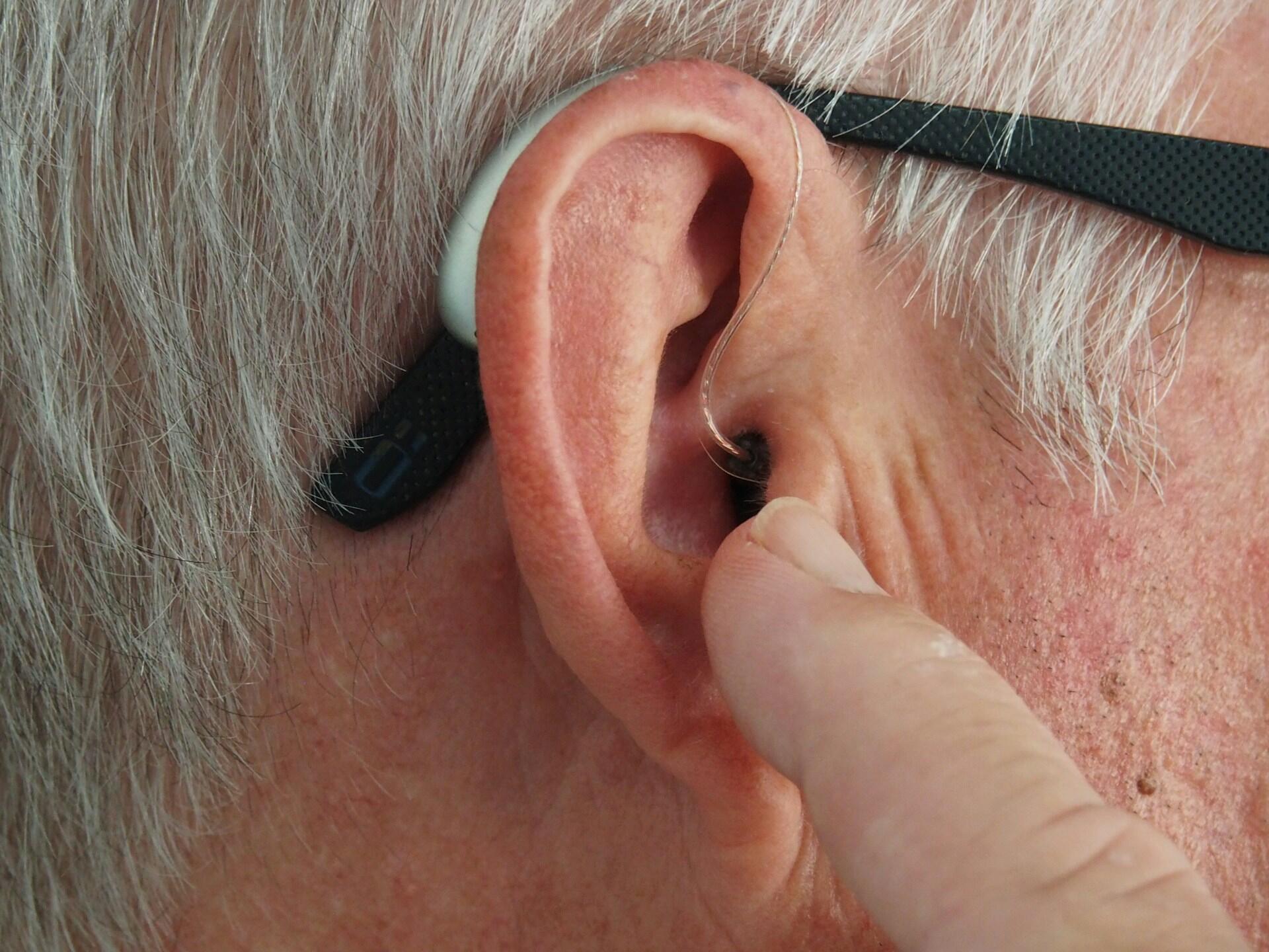 A silver-haired person points to their ear. 