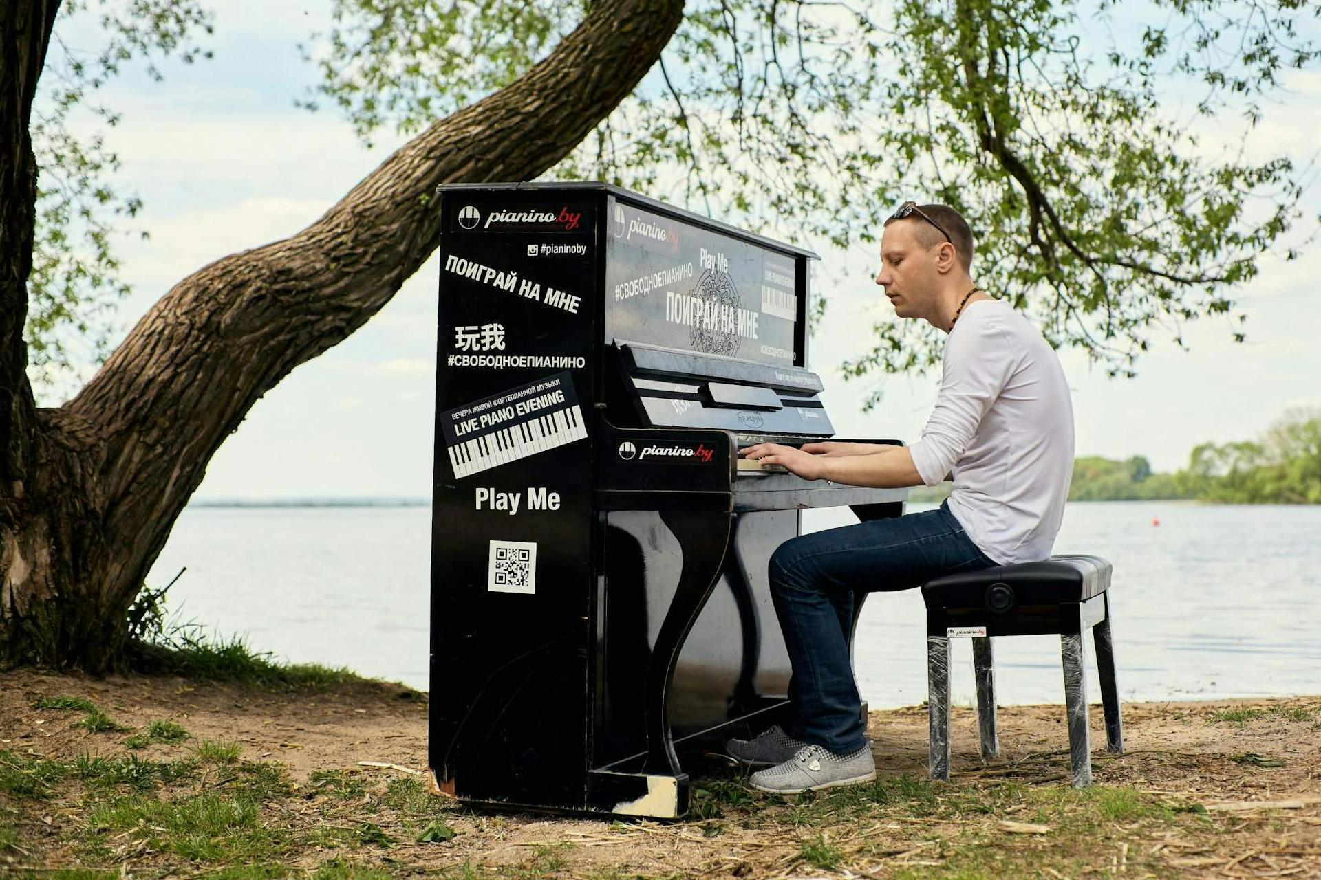 Person playing a black upright piano outdoors near a tree, with a lake in the background and "Play Me" signs on the piano.
