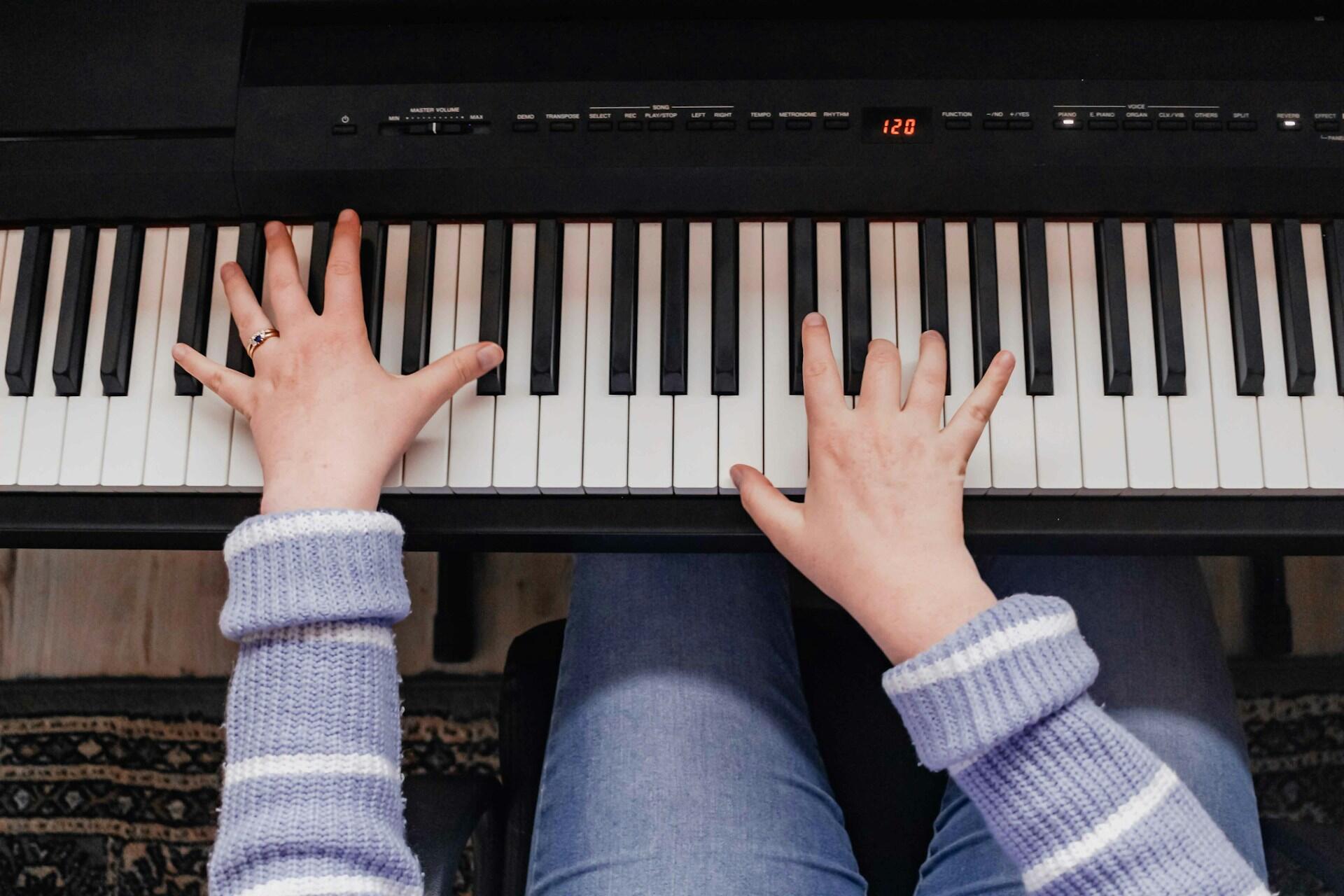 Hands of a person playing an electronic keyboard piano, wearing a purple sweater and blue jeans.
