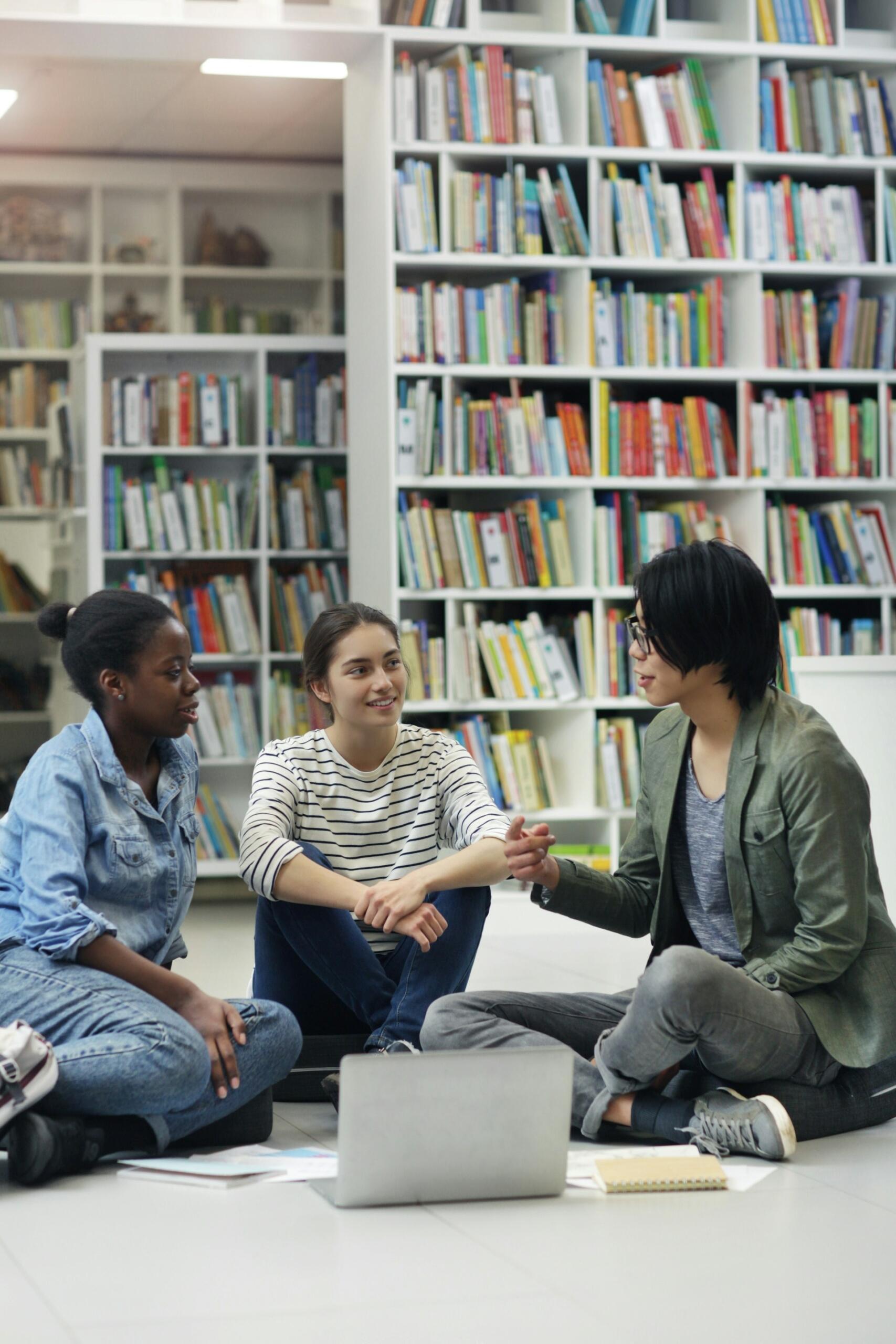 Three people sit on the floor surrounded by bookshelves. 