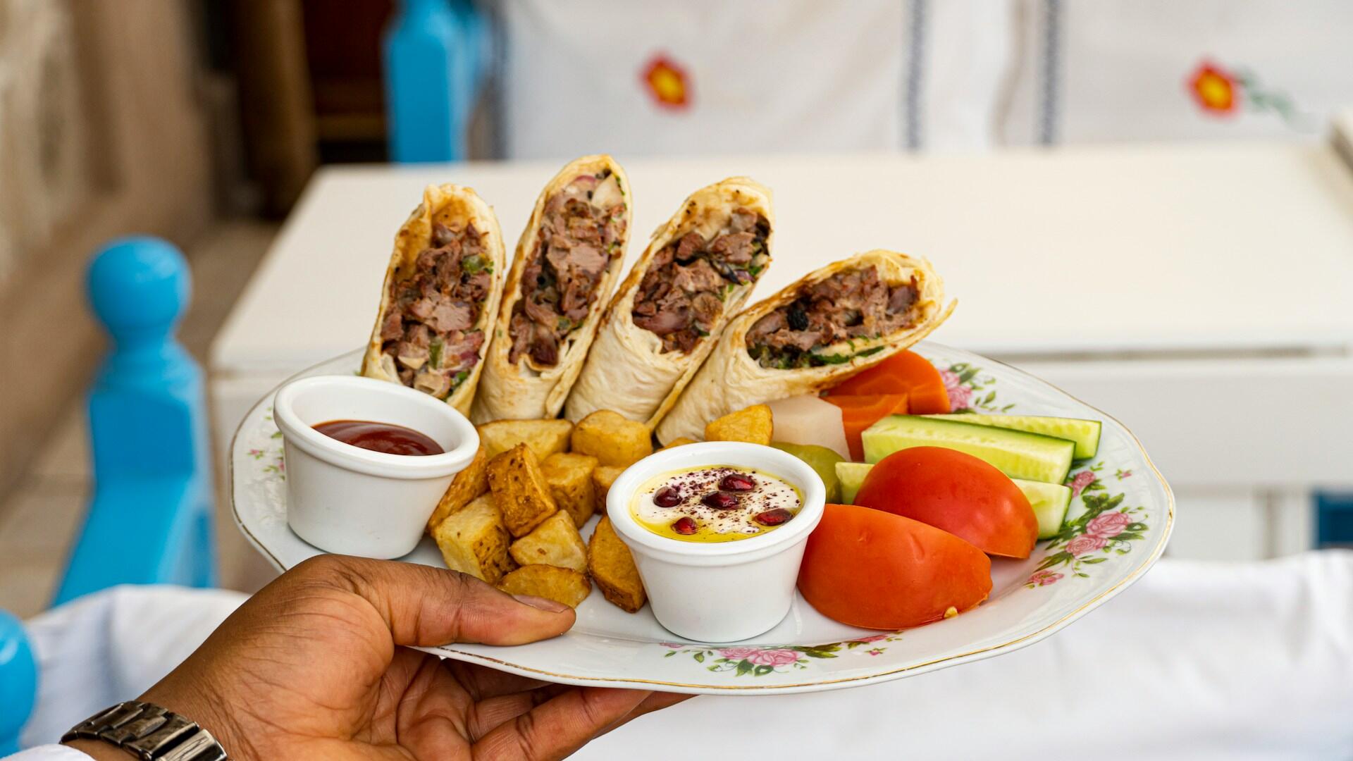 Hand holding a plate with sliced meat wraps, roasted potatoes, tomato, cucumber, carrot, and two dipping sauces.