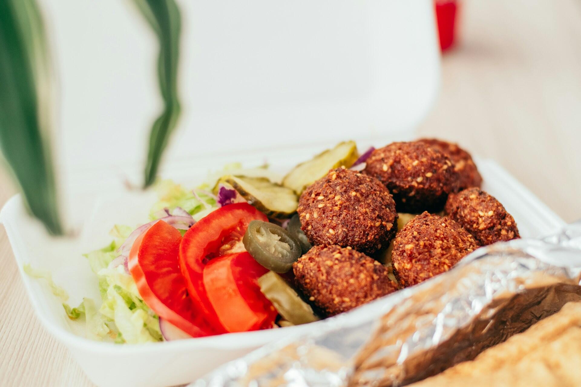 Takeout container with falafel balls, sliced tomatoes, jalapeños, pickles, and shredded lettuce on a light wooden surface.