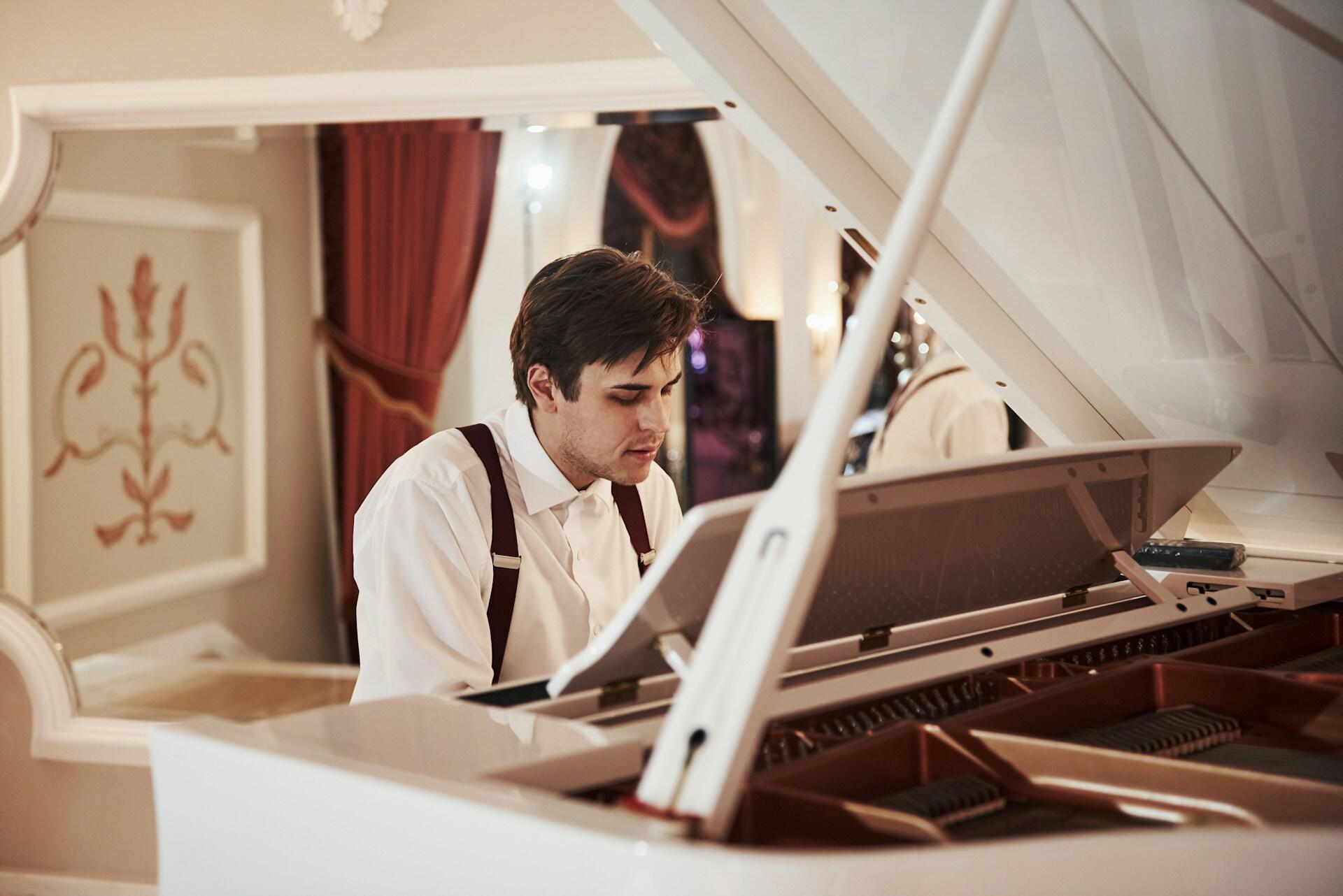 Man in white shirt and suspenders playing a white grand piano in an elegant room with red curtains and ornate decor.