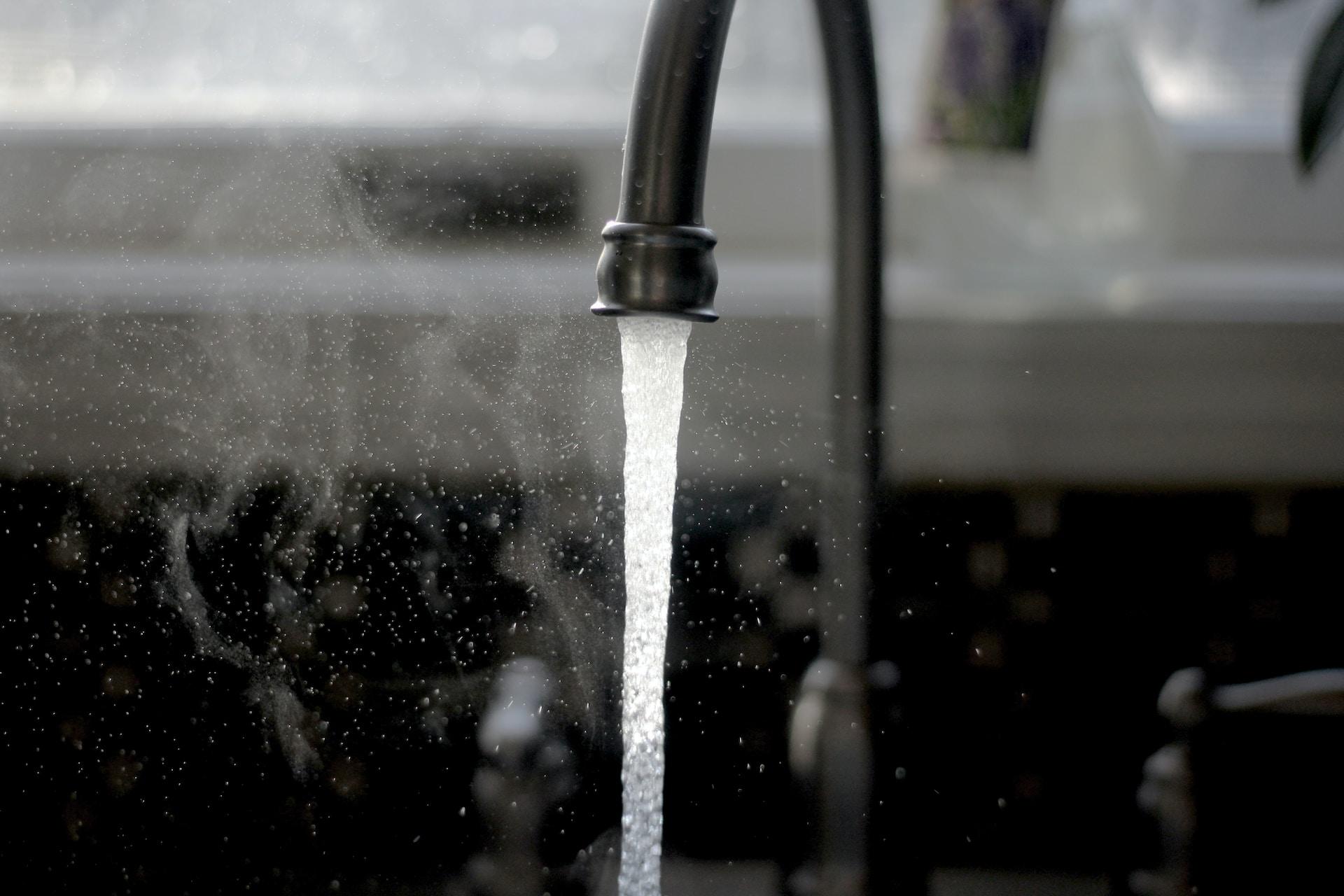 A kitchen tap with water flowing brightly in contrast to the dark splash board behind it.  