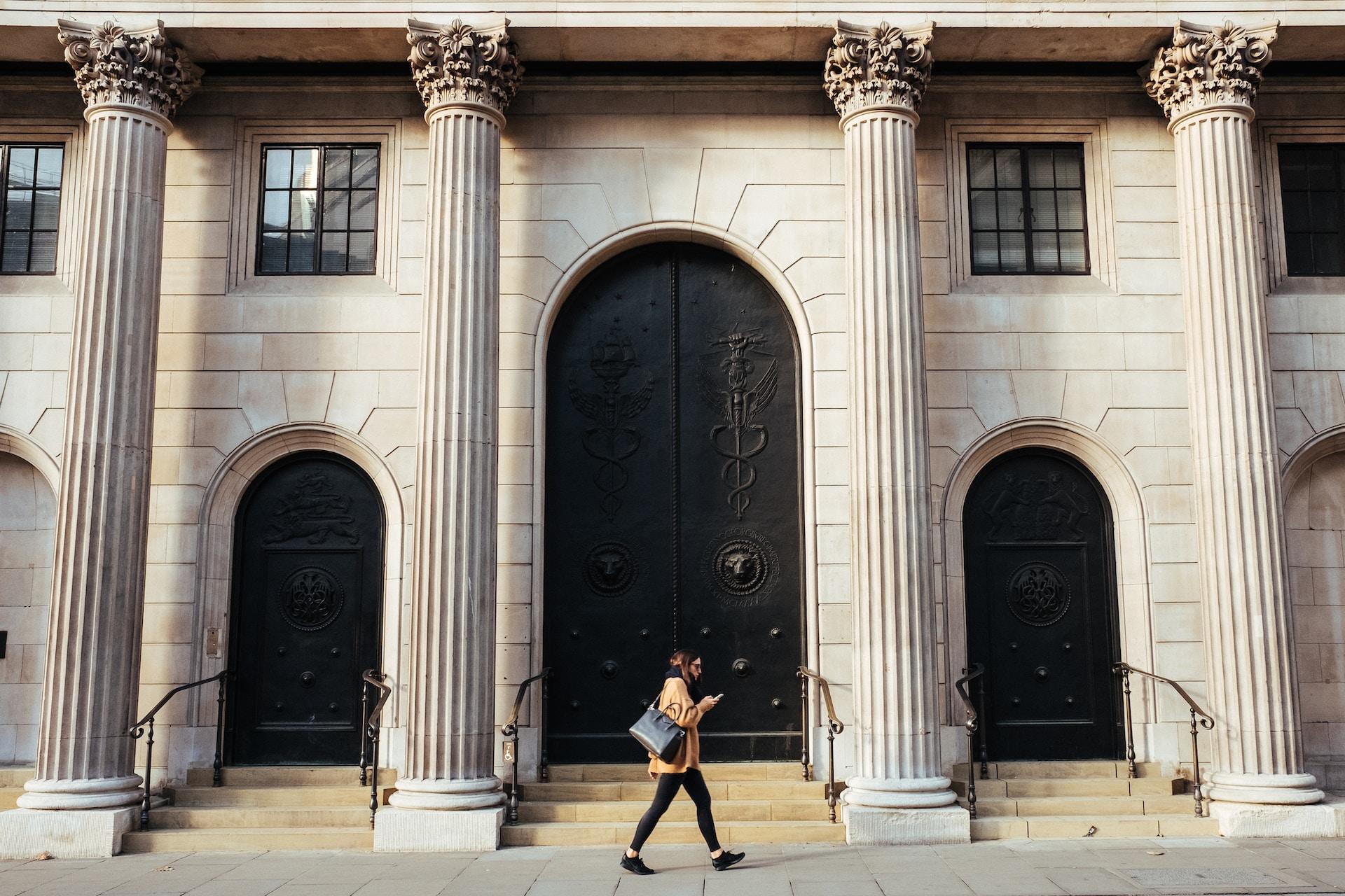 A person wearing dark lettings and a tan jacket walks past the Bank of England's main entrance, whose doors display the physicians' symbol.