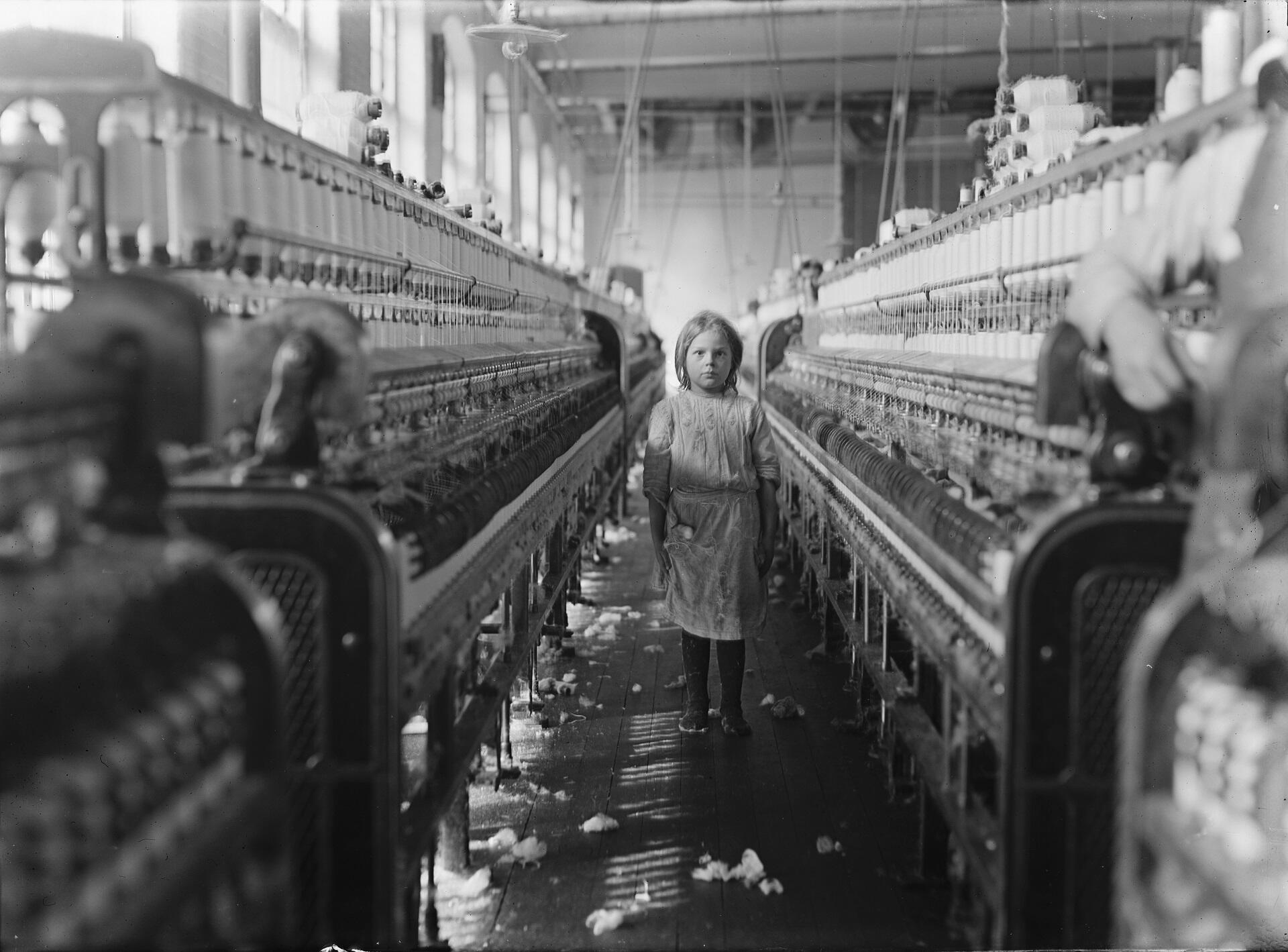 A young girl wearing a dress stands between two large industrial machines on a factory floor.
