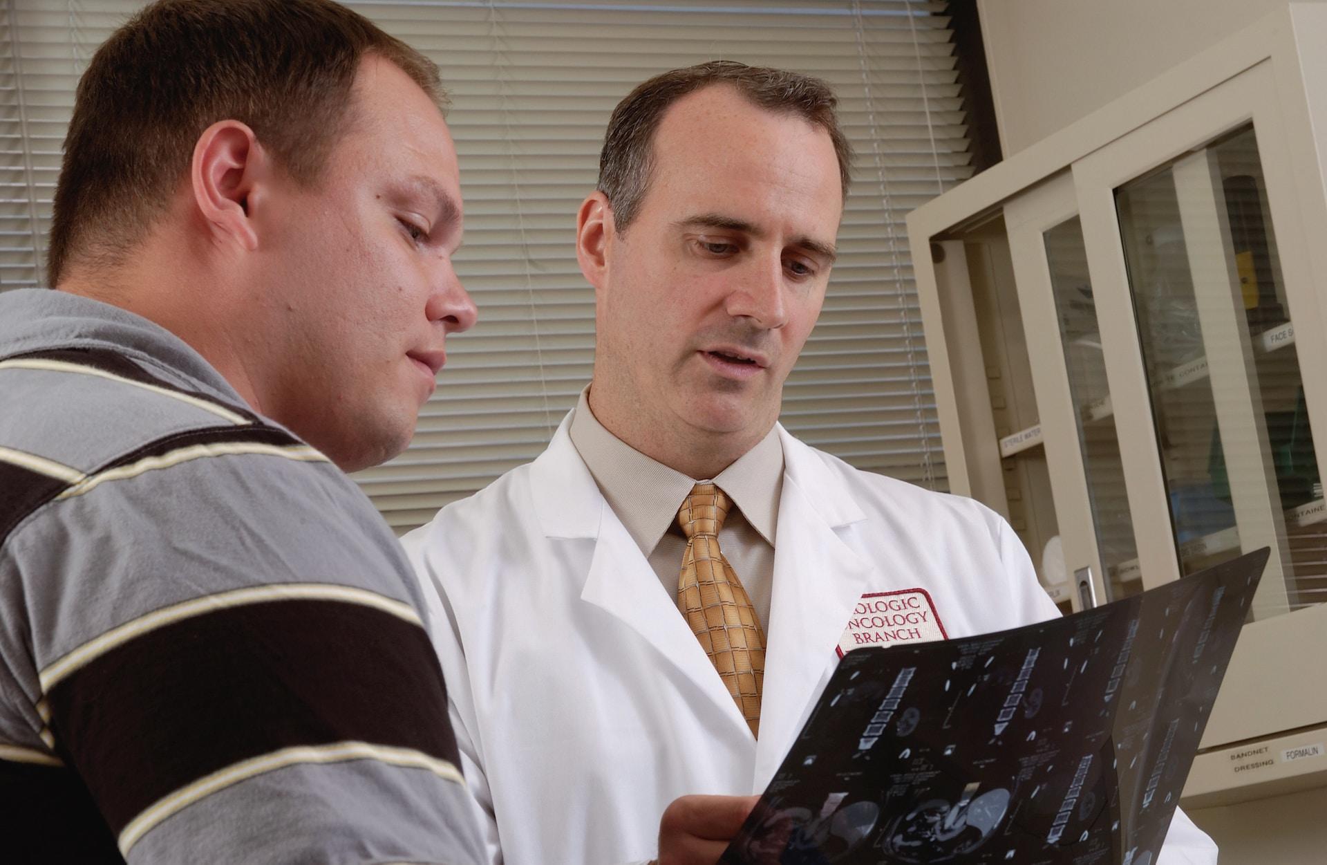 A person in a white lab coat with a red patch that says oncology research holds a screening film up and points at something on it while a patient wearing a grey, brown and white shirt looks on with a worried expression. 