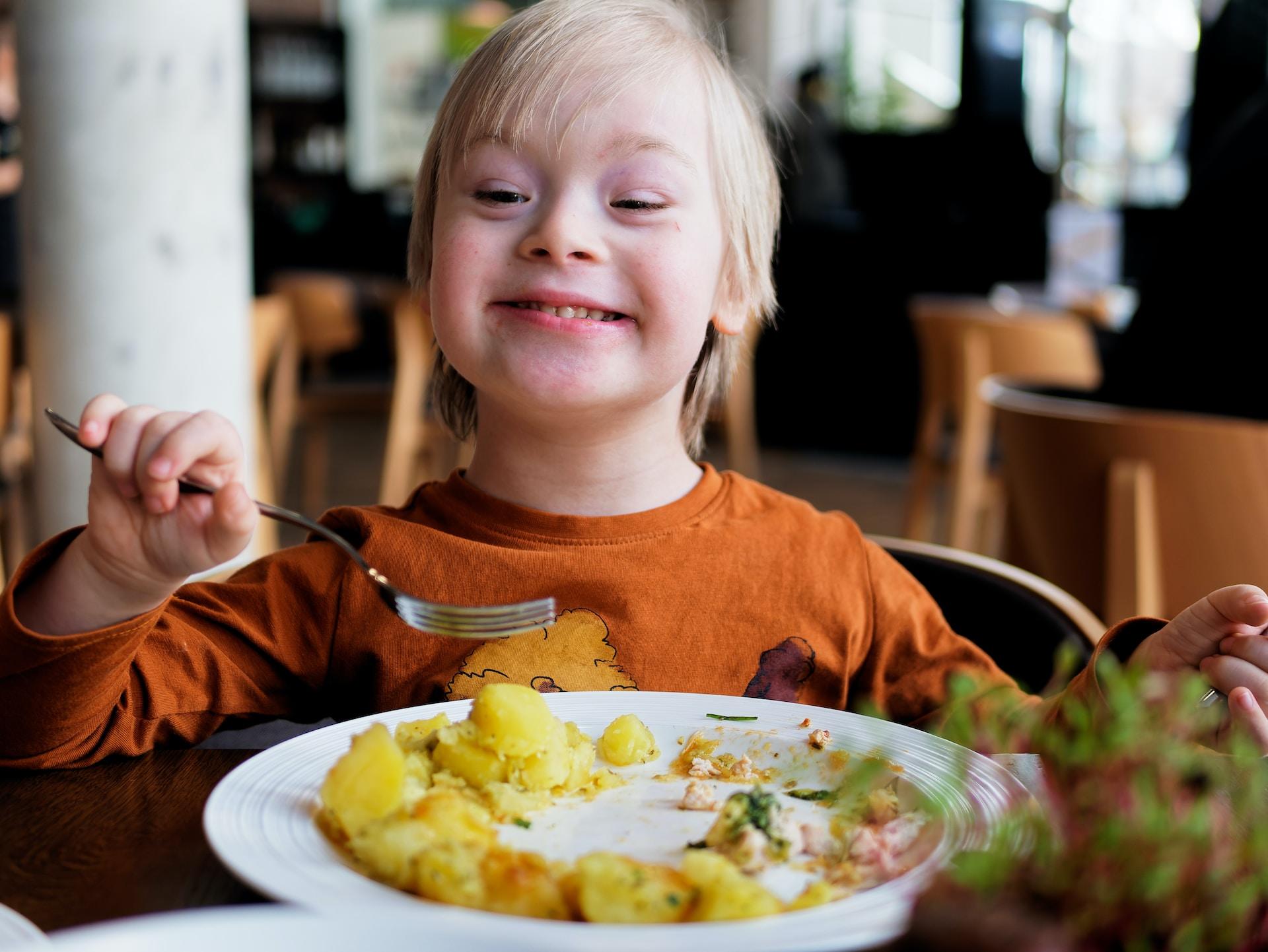 A person with blonde hair whose facial features bear signs of Down's Syndrome smiles as they hold a fork in their right hand, aimed at a plate of potatoes.