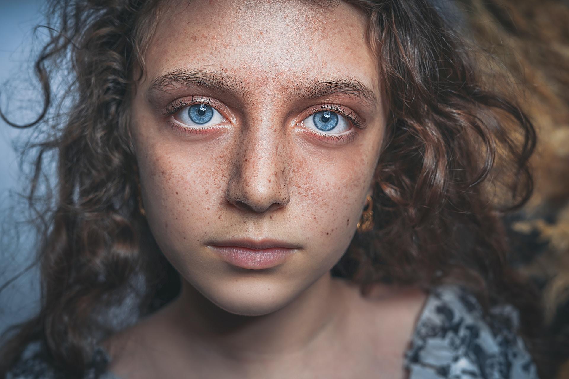 A close-up of a person with vivid blue eyes and long, wavy hair looks unsmilingly at the camera, their freckles well visible.