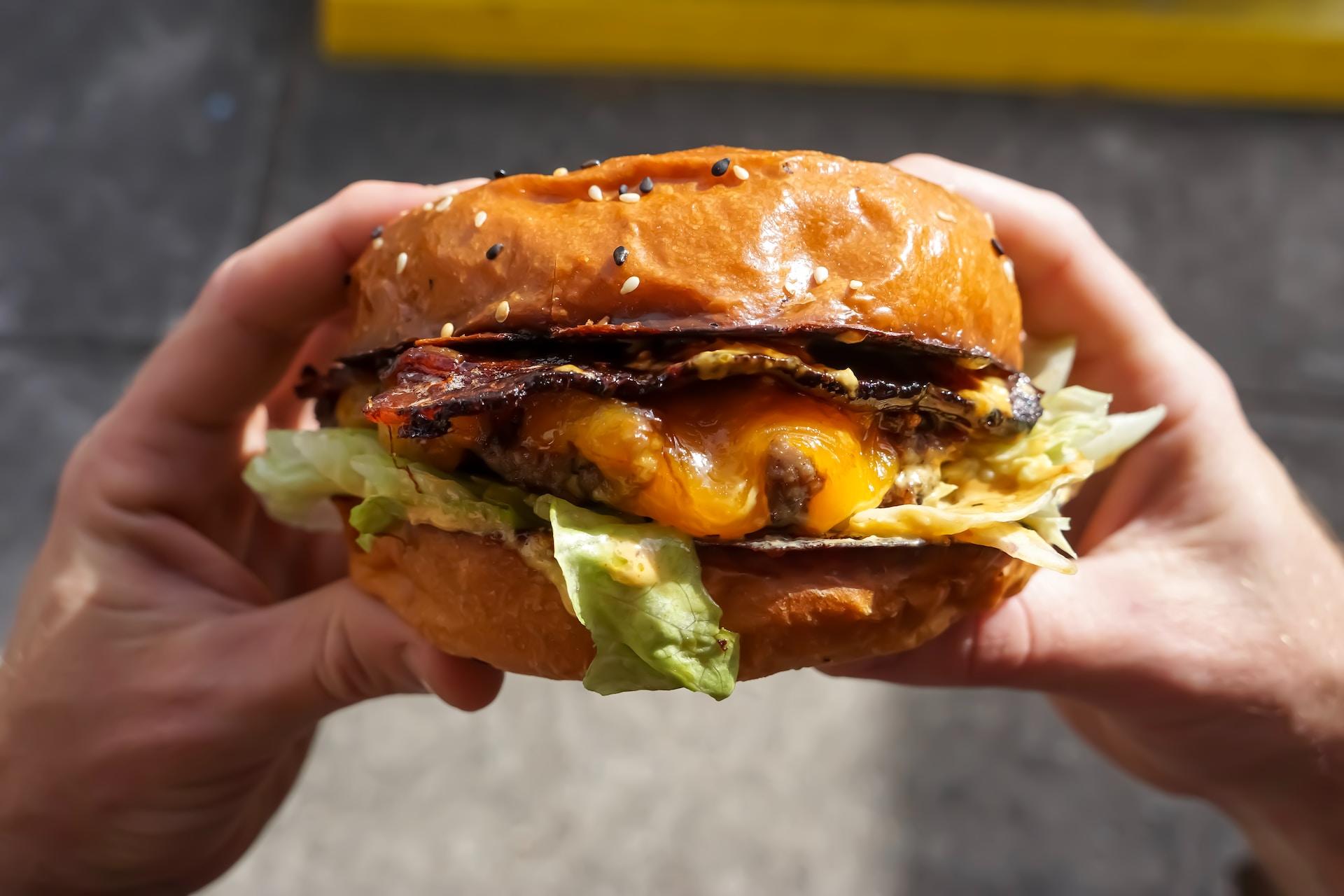 A person holds up a burger loaded with fake cheese and meat on a golden baked bun with a leaf of lettuce poking out.